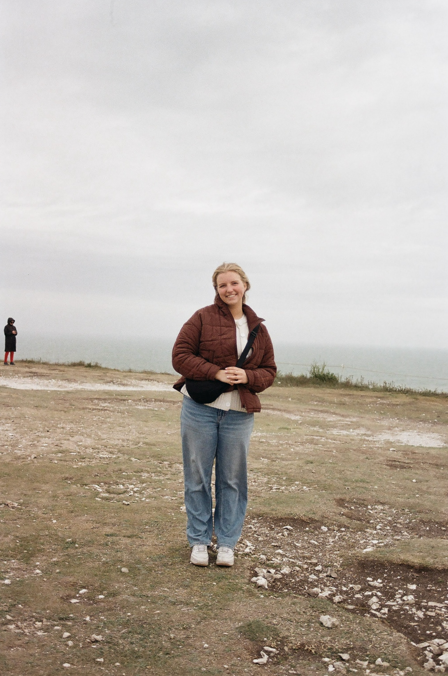 A woman with blonde hair smiling, wearing a brown jacket, white shirt, and blue jeans, standing outdoors on a cloudy day with two other people in the background.