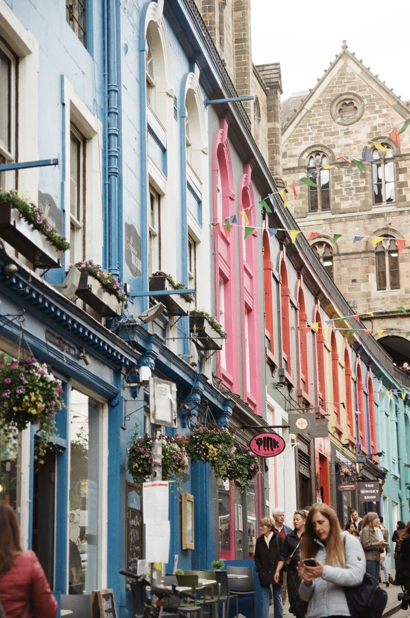 A colorful street scene with multi-colored buildings, including blue, pink, and light green, decorated with flowers and string lights. Pedestrians walk along the sidewalk, some engaging with their phones, and there are signs for businesses such as a 