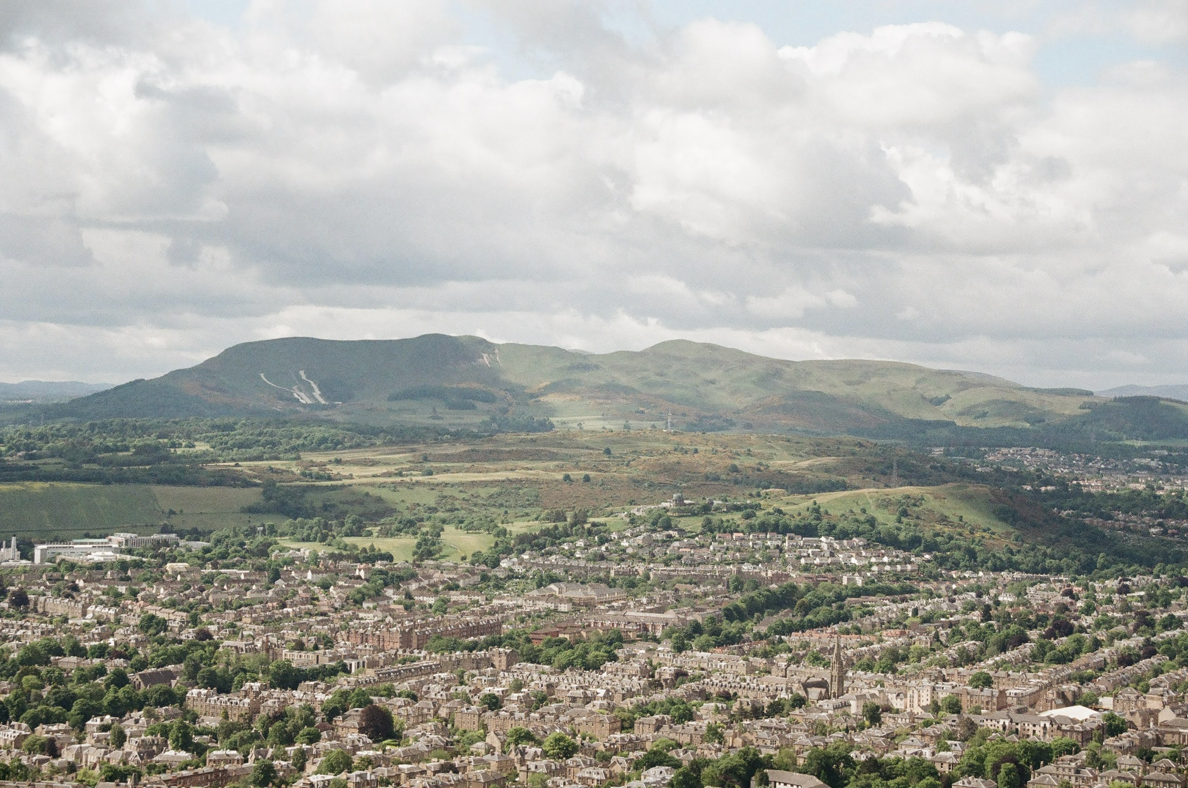 A cityscape with numerous houses and buildings in the foreground, followed by green fields and rolling hills, and mountains in the background under a partly cloudy sky.