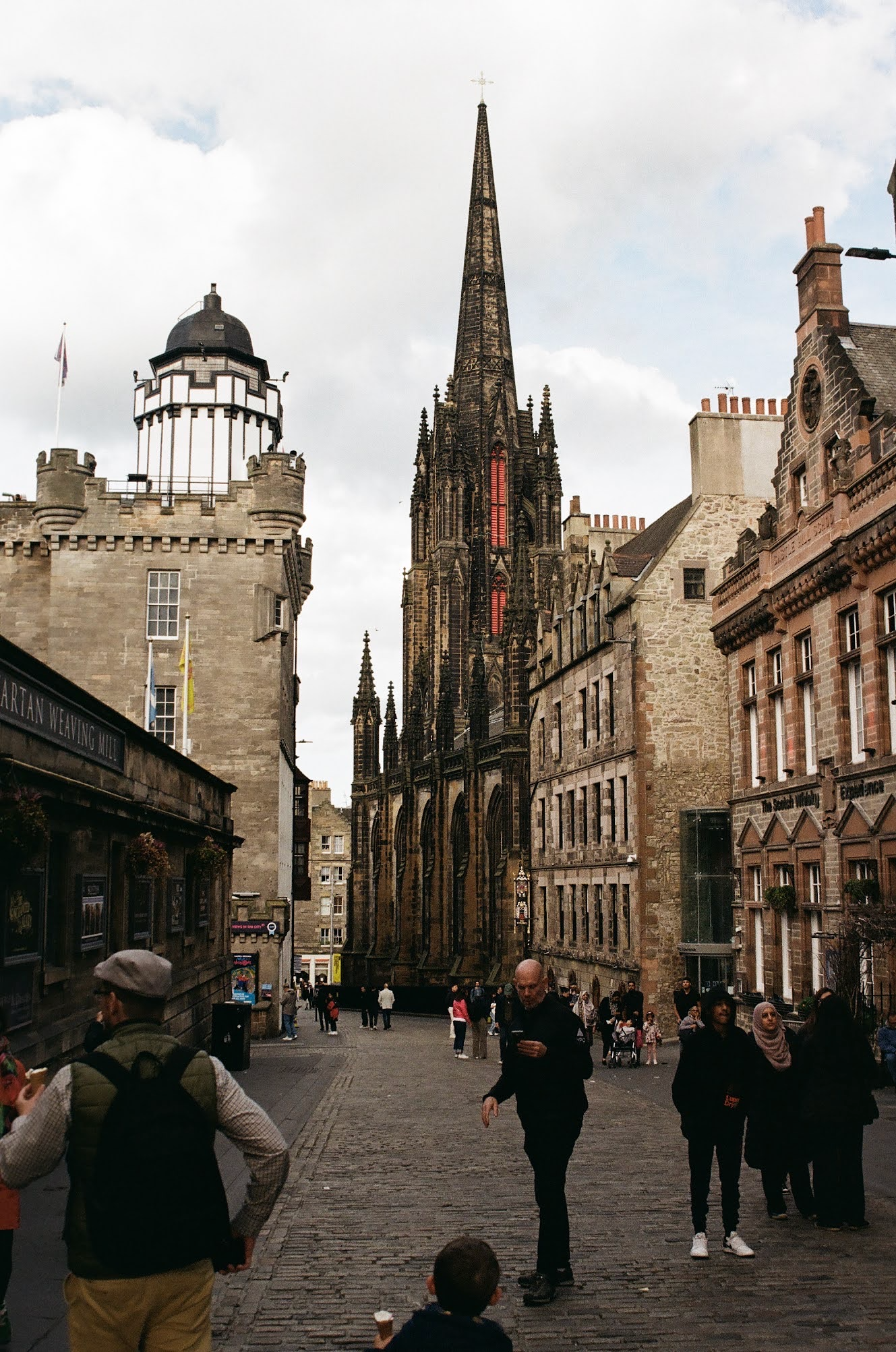Crowded street scene in front of a historic Gothic church with a tall spire, surrounded by old stone buildings and people walking or standing, some with shopping bags or taking photos.