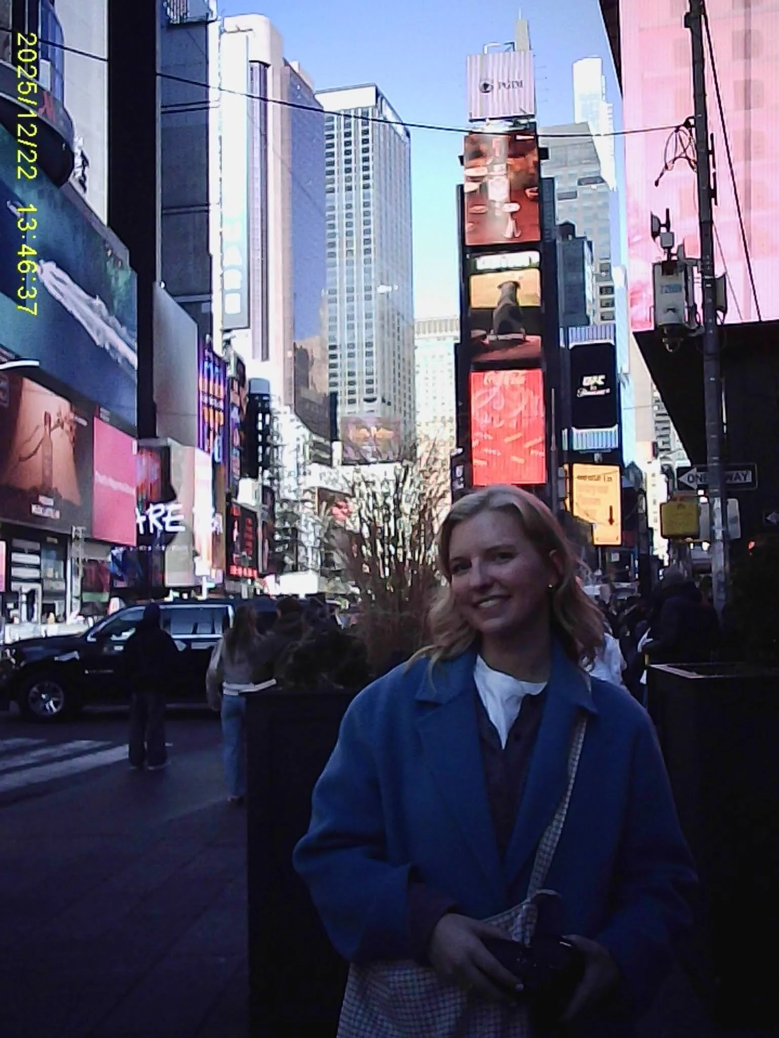 A woman with shoulder-length hair smiling in Times Square with illuminated billboards and a crowd of people behind her.