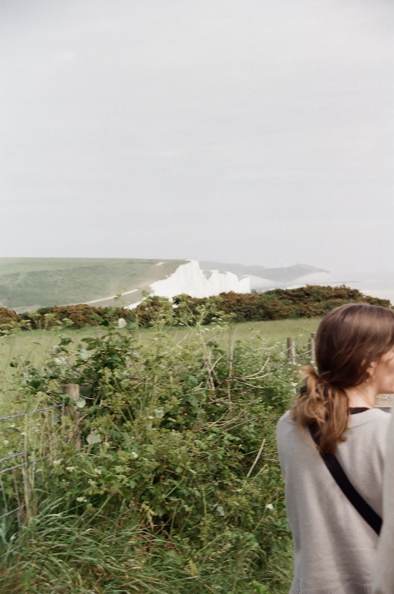A woman with brown hair in a ponytail and a light-colored sweater stands in front of a green field with shrubs and a wooden fence. In the background, there are white chalk cliffs and a cloudy sky.