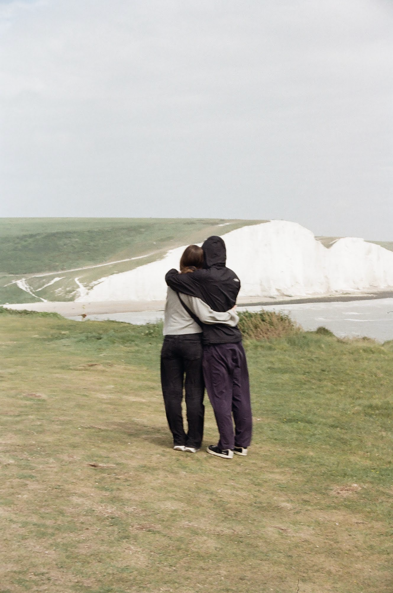 Two people, one in a black jacket and the other in a gray and black jacket, hugging outdoors on a grassy area near white chalk cliffs with green slopes in the background.