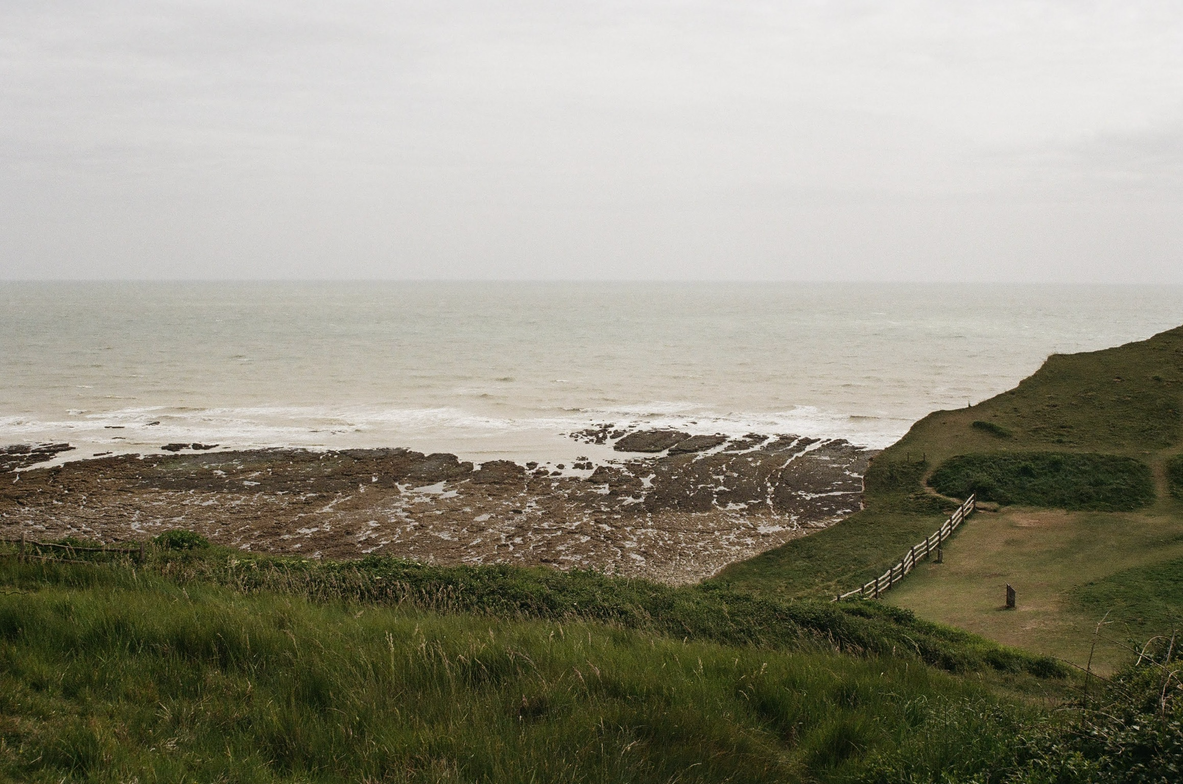 A coastal scene with green grassy dunes in the foreground, a rocky shoreline and ocean waves in the midground, and a cloudy sky overhead.