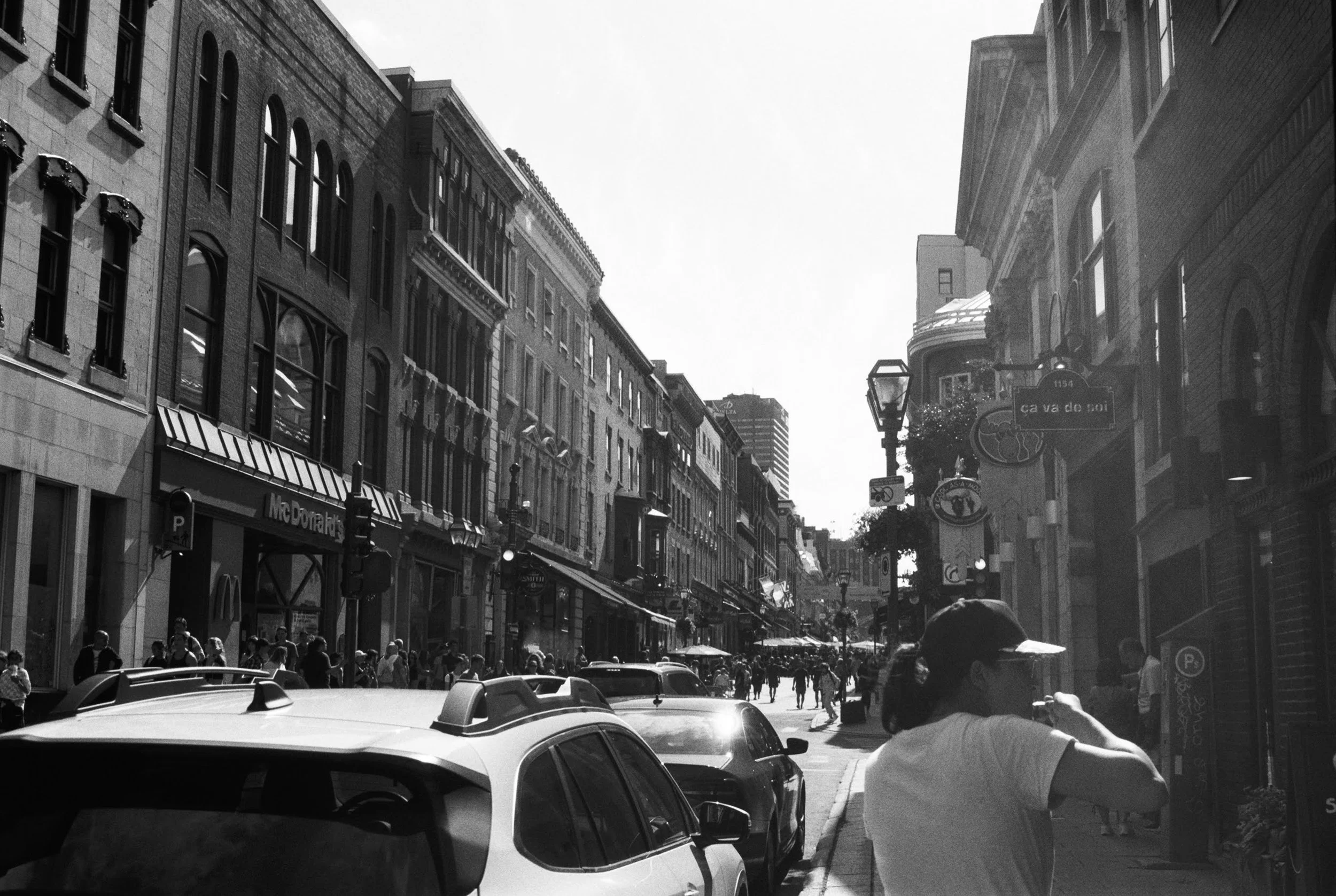 A busy city street scene in black and white with people walking on the sidewalk, cars parked and driving, multi-story buildings with signs and awnings, street lamps, and a street name sign reading 'Cava de'.