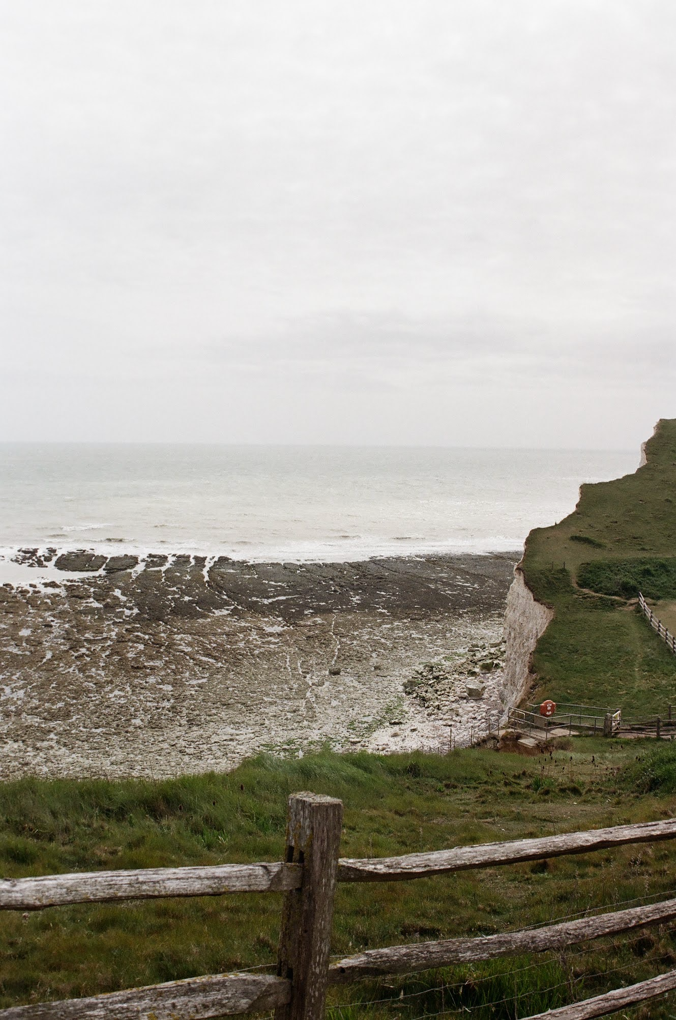 Cliffs overlooking a beach and ocean, with a grassy fenced area in the foreground, cloudy sky.