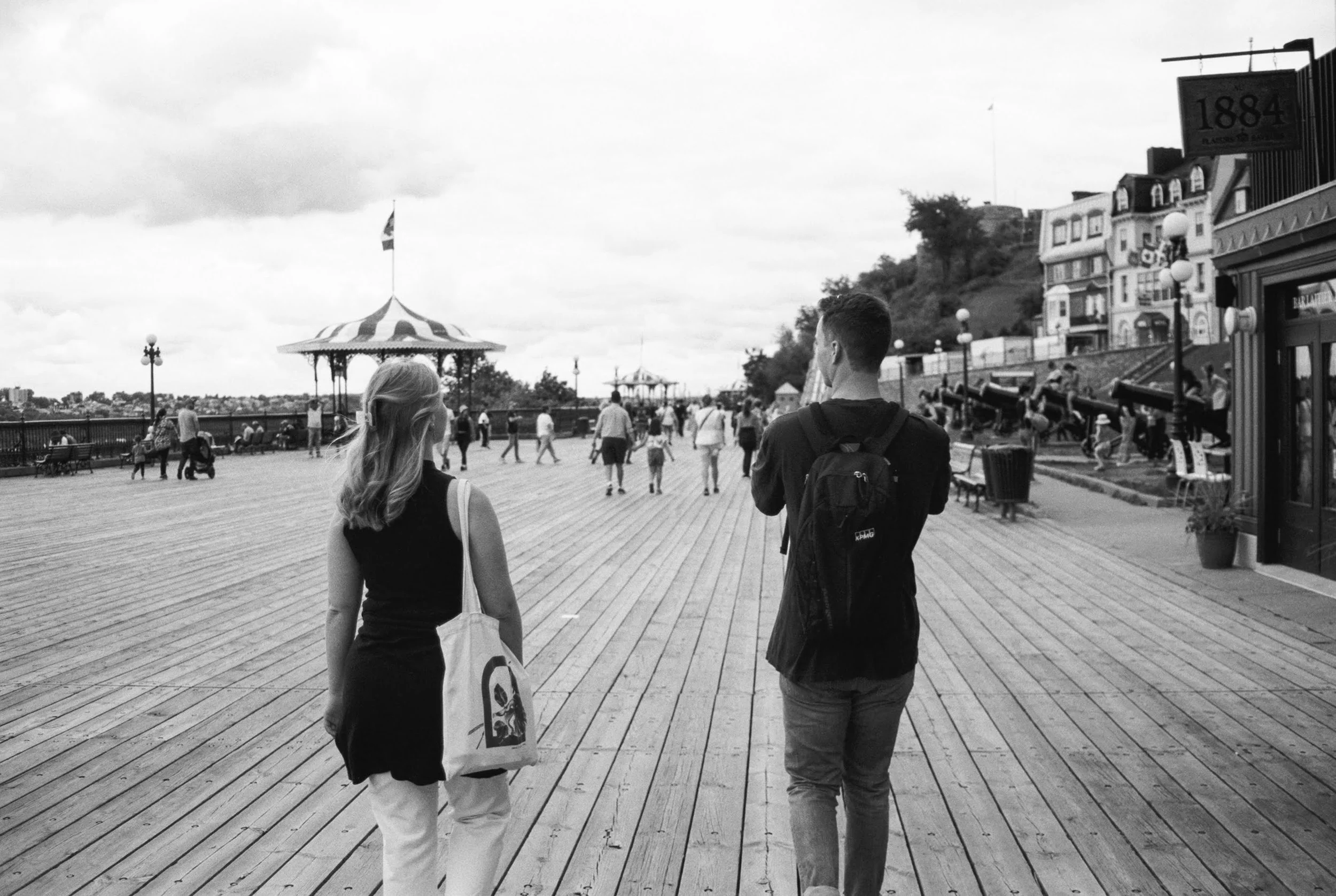 People walking on a wooden boardwalk in a seaside town, with buildings, a carousel, and a hill in the background in black and white.