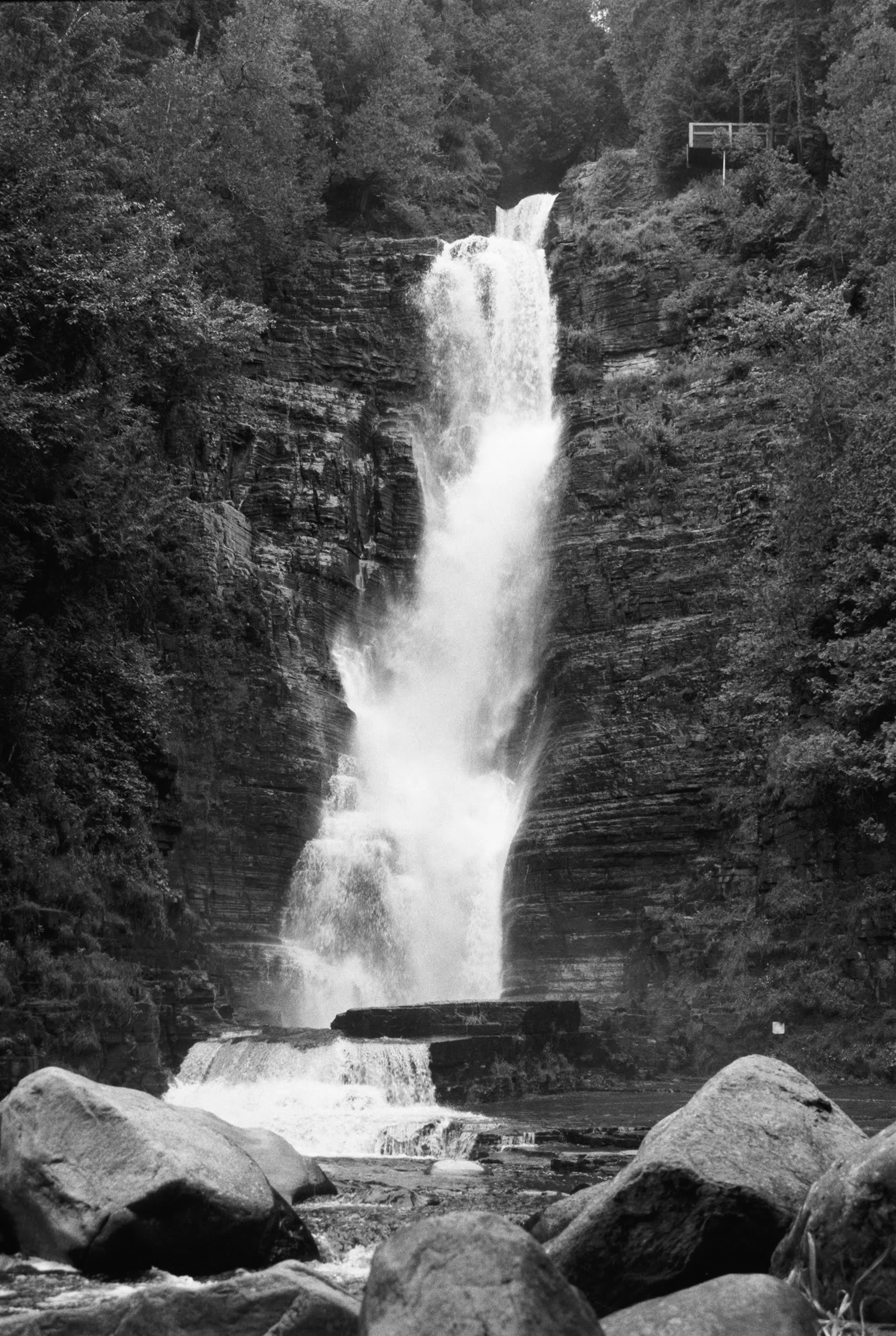 A black and white photo of a waterfall flowing down a rocky cliff surrounded by trees, with large rocks in the foreground and a small platform with a railing on the right side of the top of the waterfall.