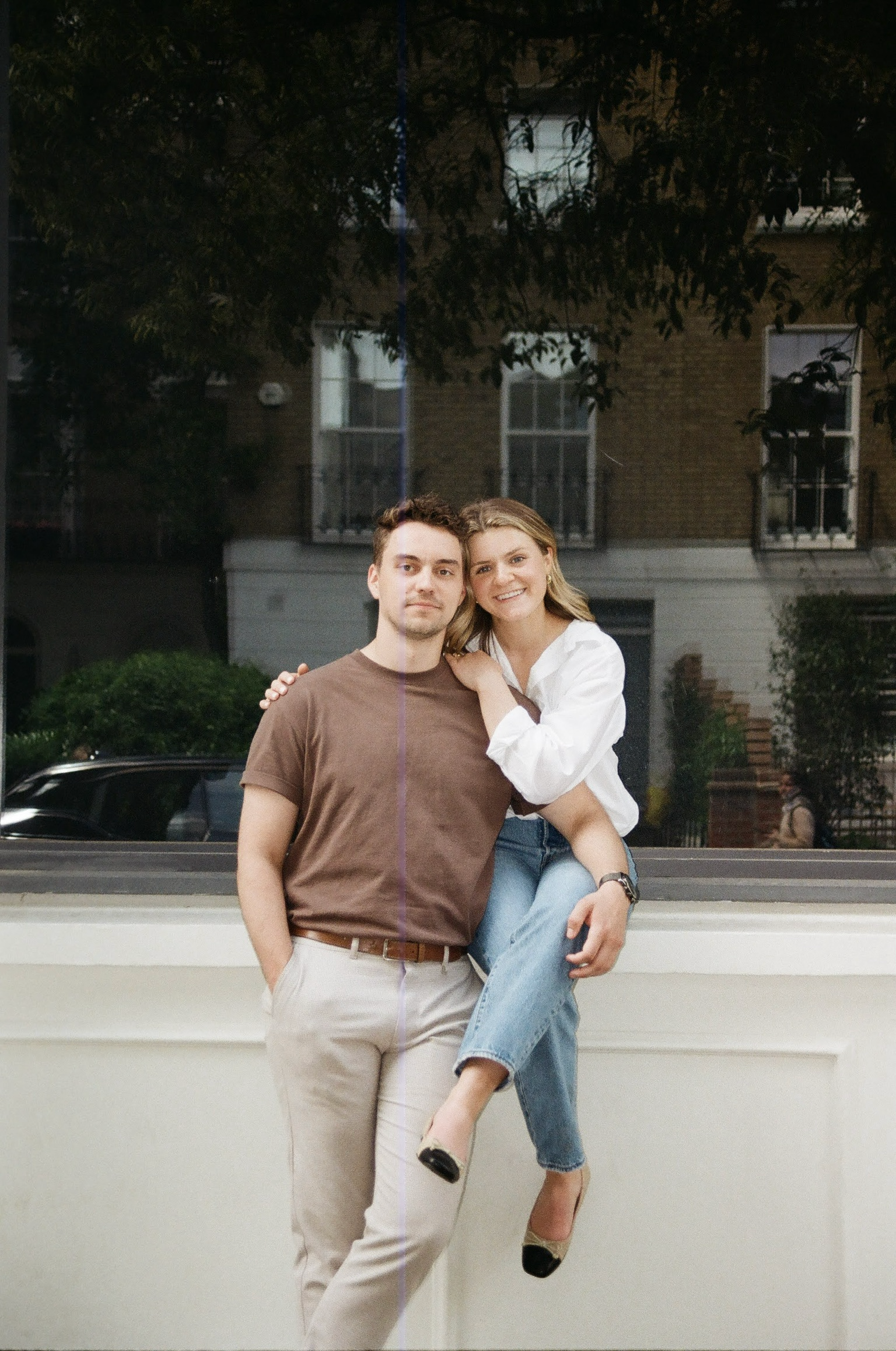A young man in a brown T-shirt and beige pants stands with a young woman in a white blouse and blue jeans, who is sitting on his lap with her arms around his shoulders, smiling, in front of a city building at night.