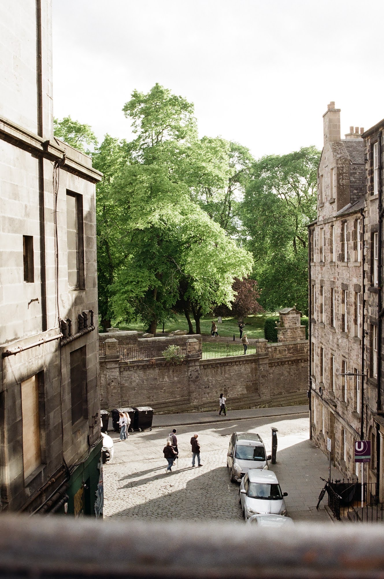 Urban view with cobblestone street, parked cars, people walking, stone and brick buildings, and a park with green trees in the background.
