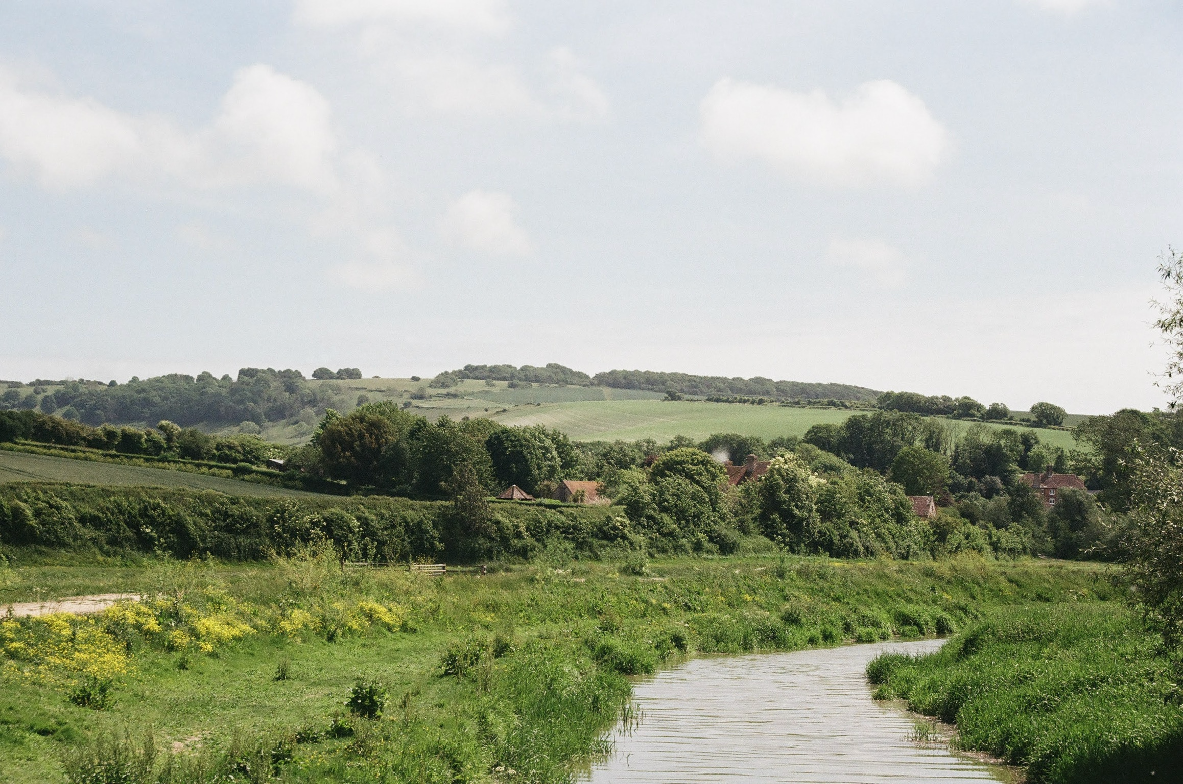 A landscape of green rolling hills with a river flowing through the foreground, trees scattered across the hills, and a partly cloudy sky overhead.