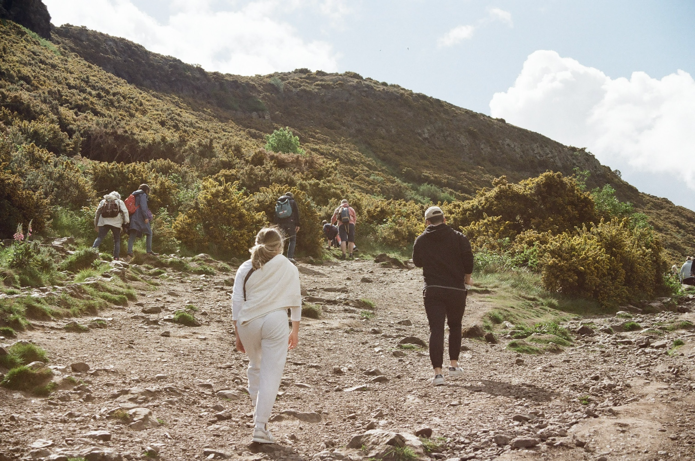 People hiking on a rocky trail through a mountainous landscape with green shrubs and trees, blue sky, and clouds.