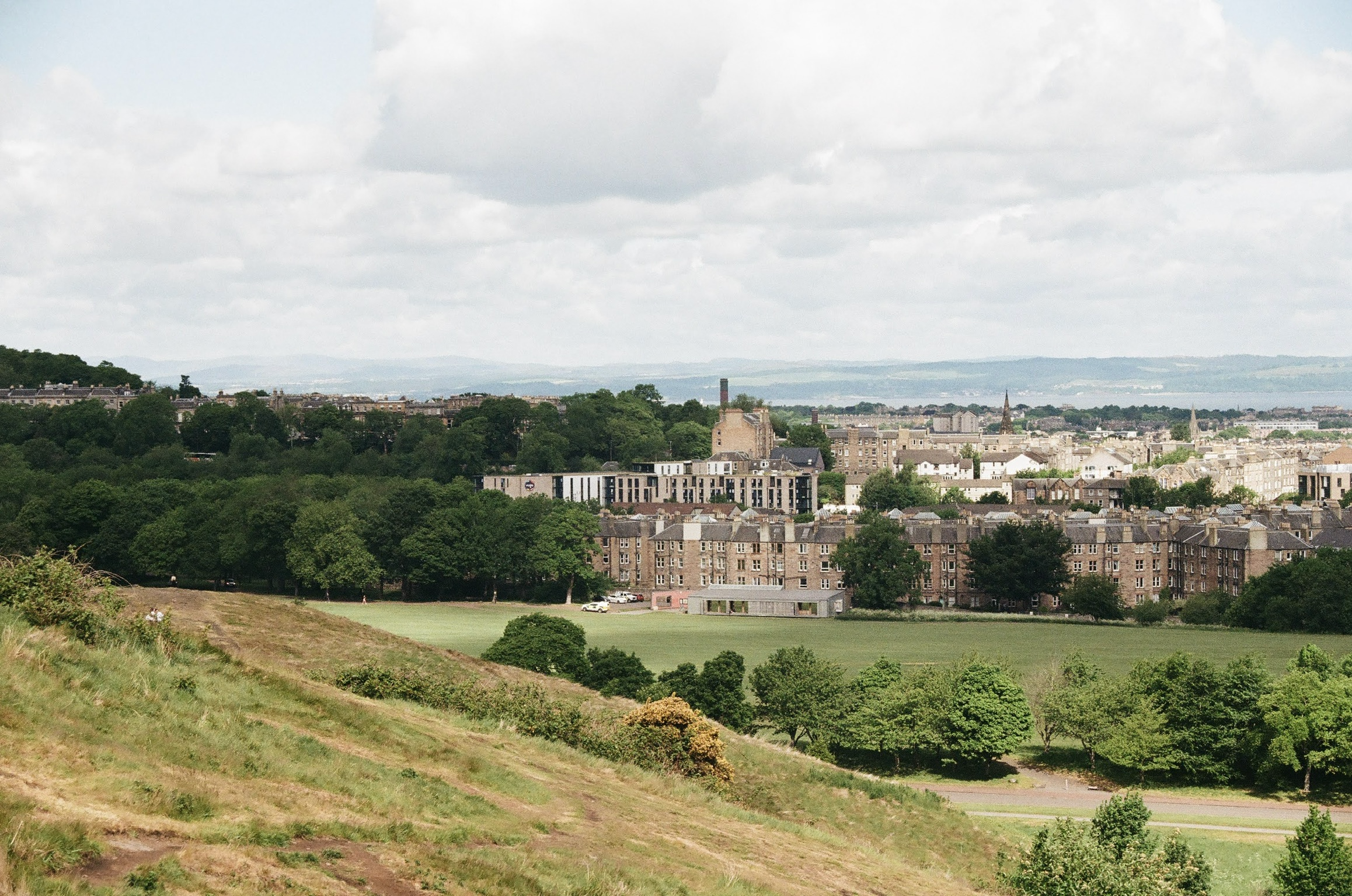 A scenic view of a city with a mixture of trees, residential buildings, and distant hills, under a cloudy sky.