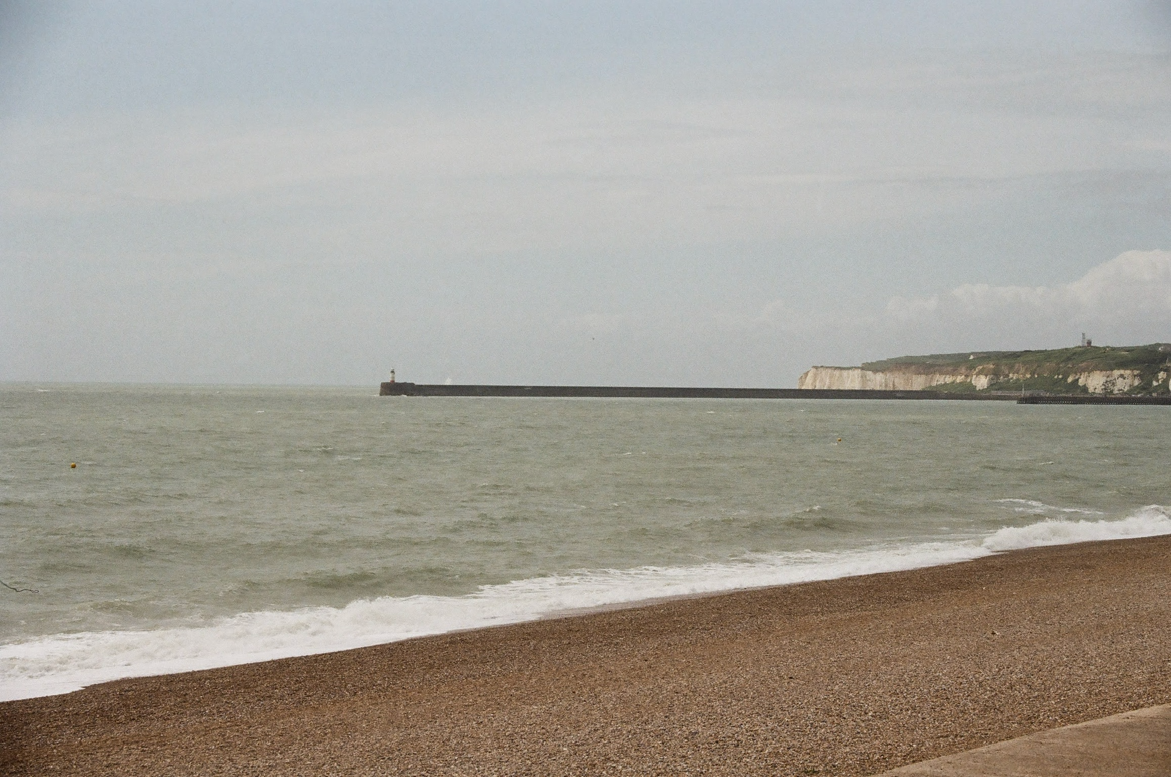 Seaside view of a sandy beach with waves and a breakwater extending into the ocean. Cliffs with greenery and structures are visible in the distance under a cloudy sky.