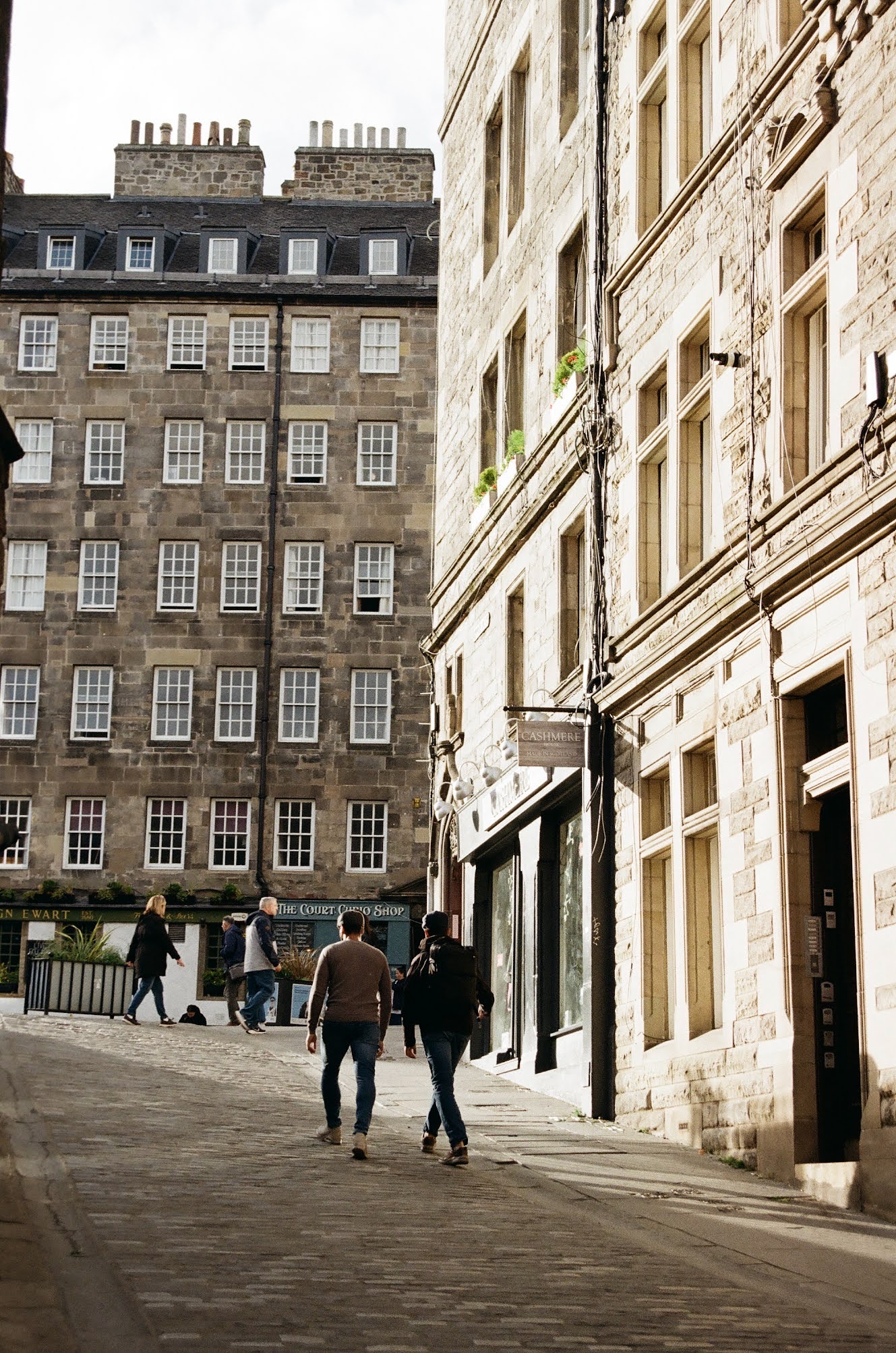 People walking along a city street with historic stone buildings and shops, including a sign that says 'Cashmere' and 'The Court Oslo Shop'.