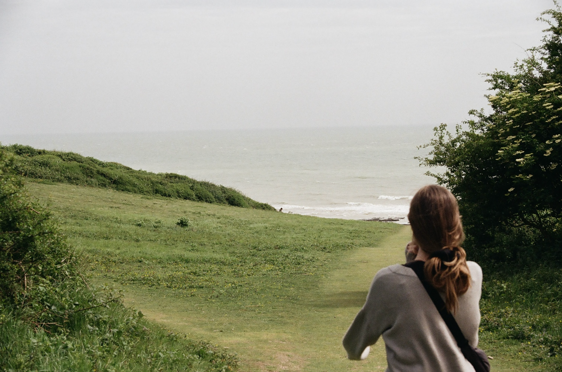 Person with brown hair running in a park by the seaside, green grass, bushes, and trees, with ocean and cloudy sky in the background.