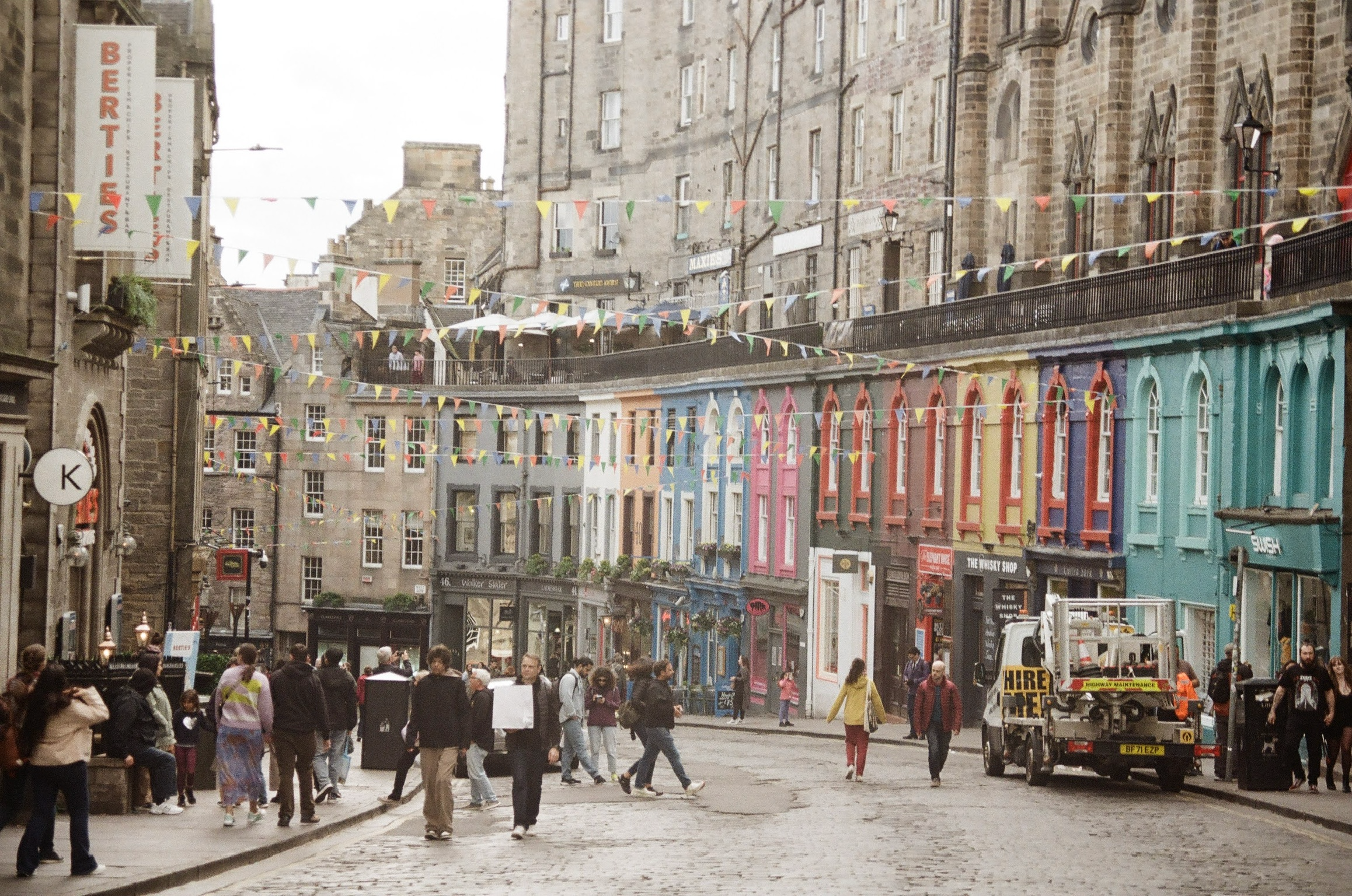 A lively city street decorated with colorful triangular banners hanging across from buildings. Crowds of people walk along the sidewalks and cobblestone street, with various shops, including a pub and a colorful row of buildings. Some construction wo
