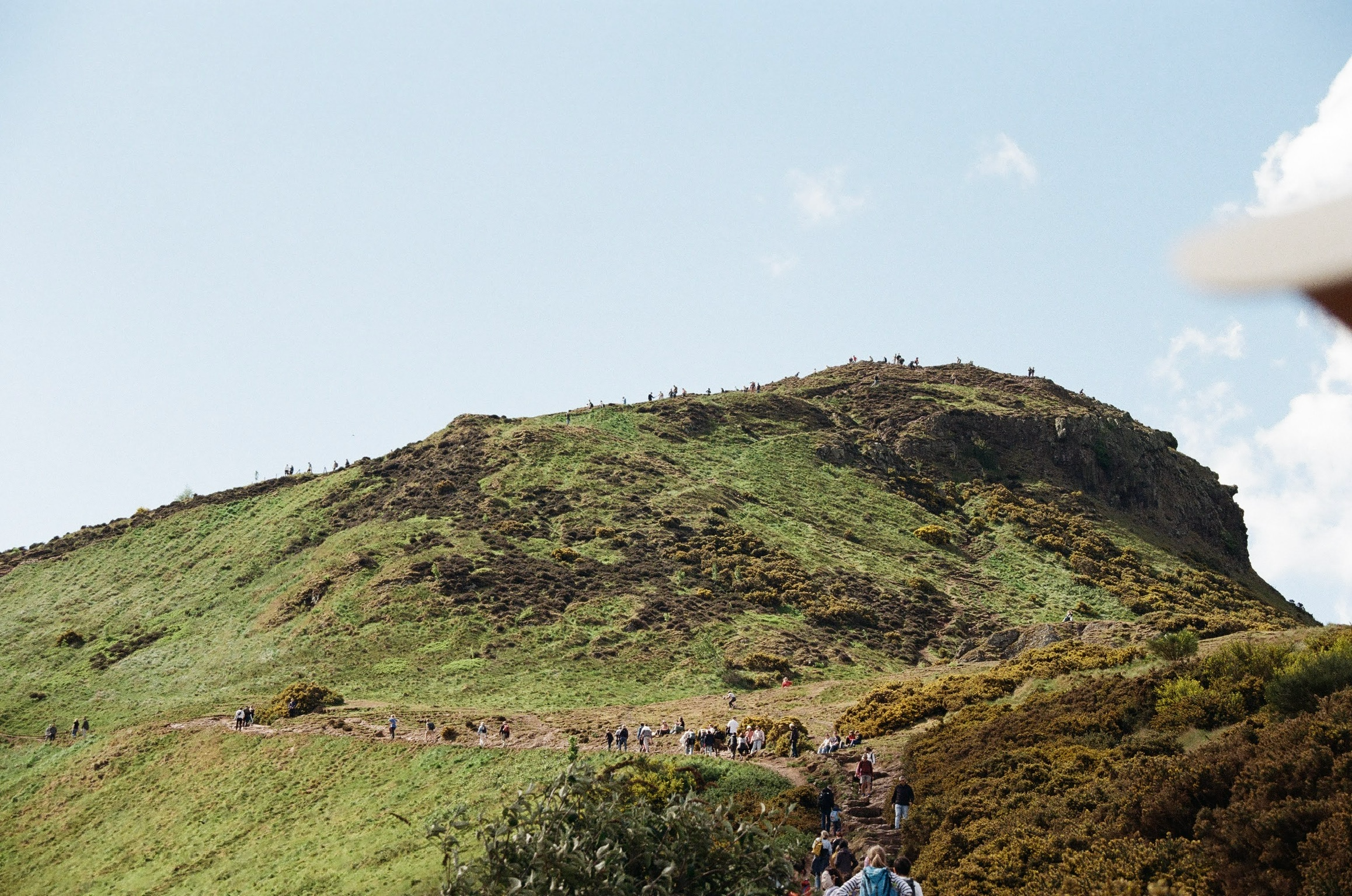 Hikers walking up a green, hilly mountain with a small crowd at the summit against a partly cloudy sky.