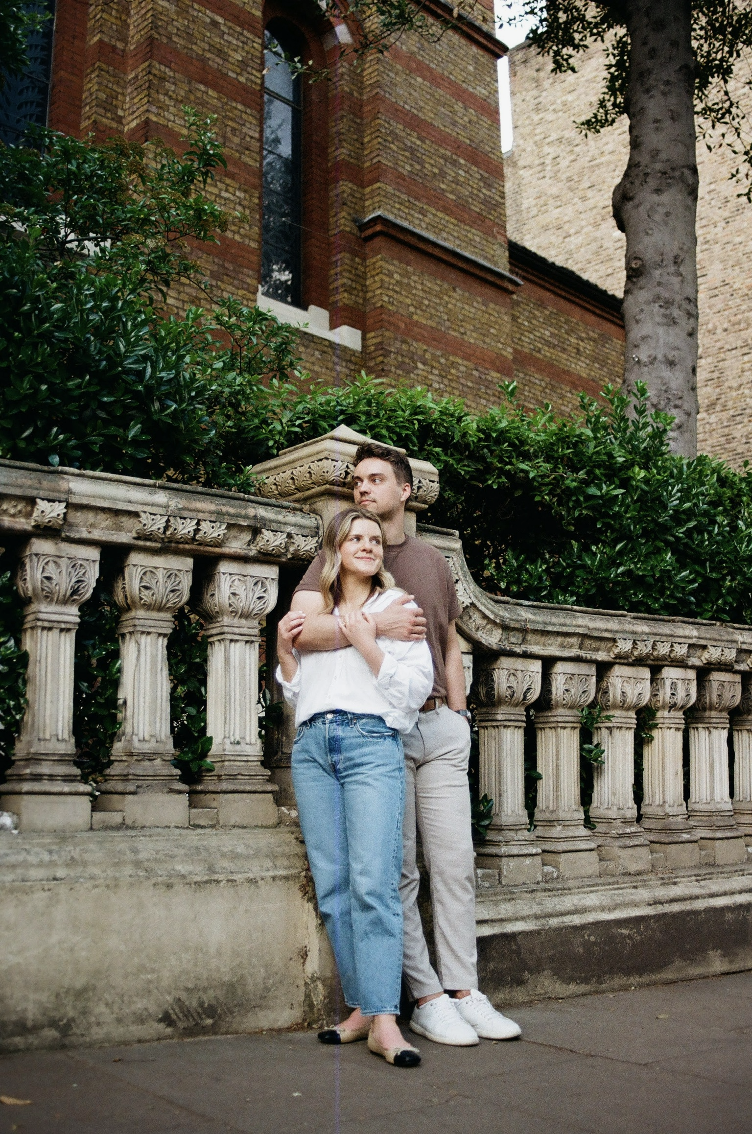 A young couple embracing on a stone staircase in front of a decorative stone railing, with lush green bushes and trees, a brick building with arched windows, and a brick wall in the background.
