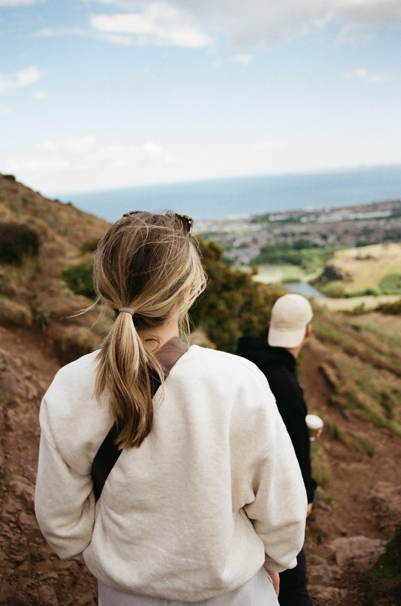 A woman with long, light brown hair tied in a ponytail wearing a white sweatshirt is walking on a dirt trail in a hilly landscape, with a person wearing a white hat and black jacket nearby, all overlooking a body of water and a distant coastline unde