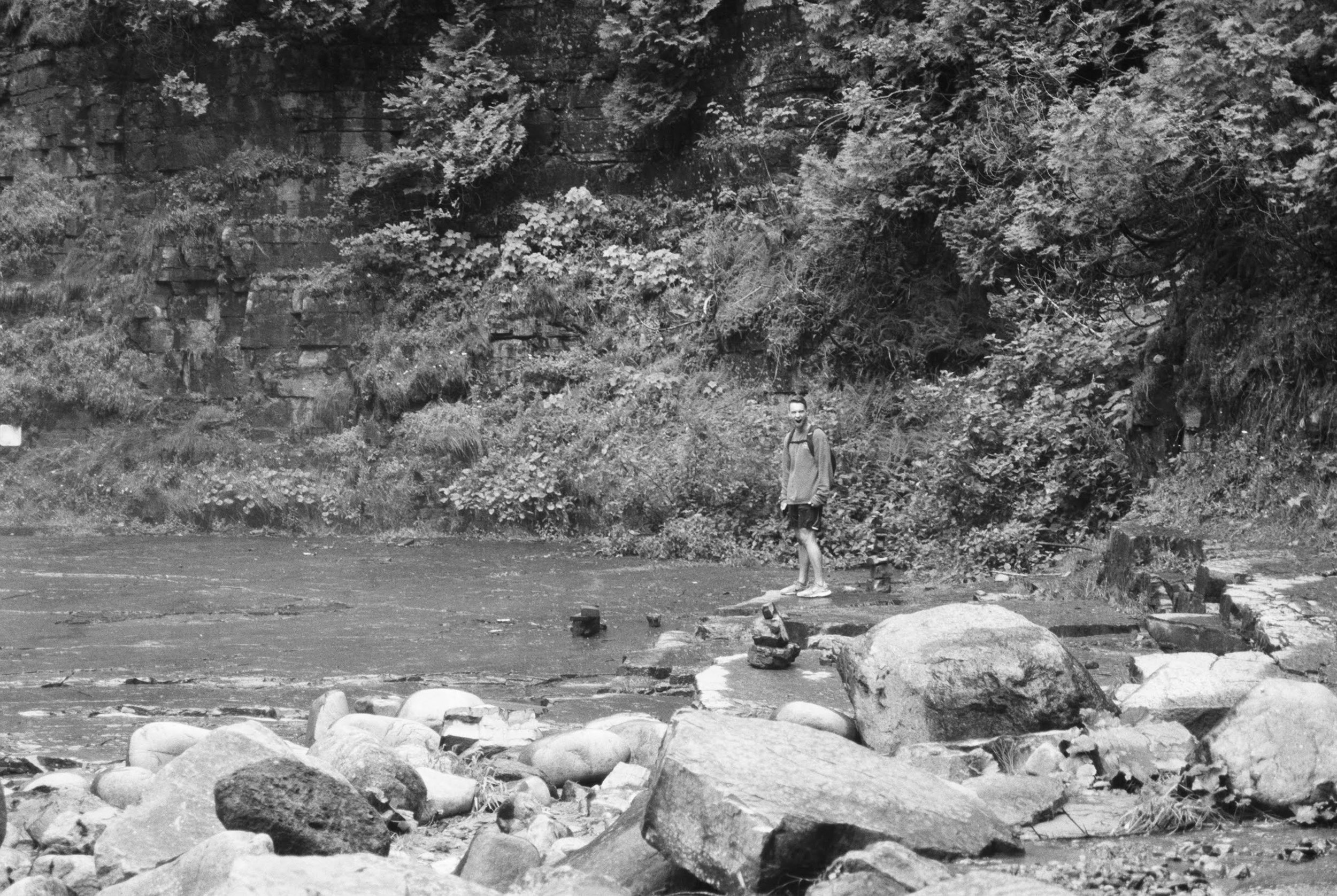 A person standing on rocks near a river with a forested hillside in the background.