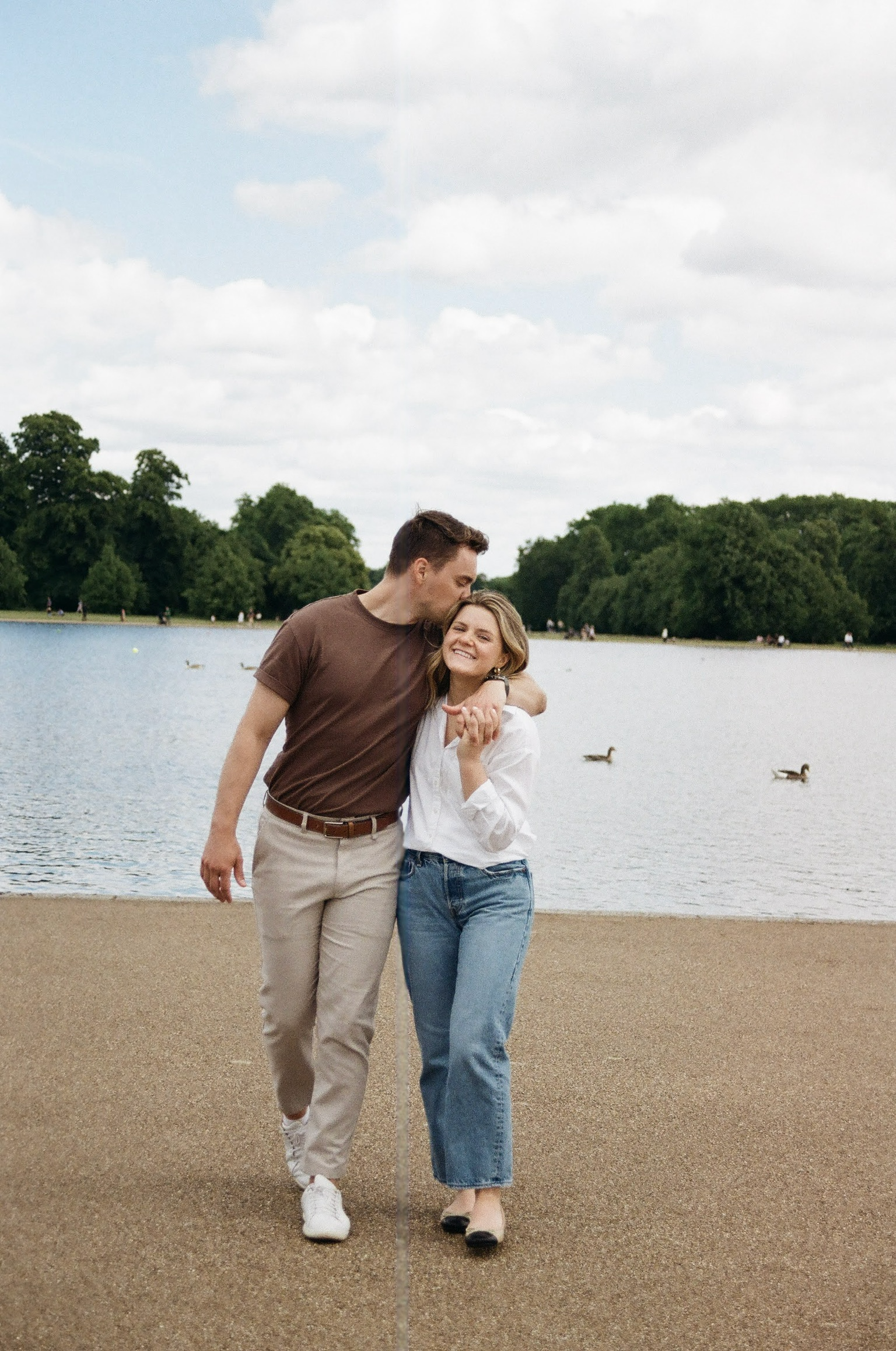A smiling woman and a man walking by a lake, with trees and ducks in the background, holding hands and embracing each other.