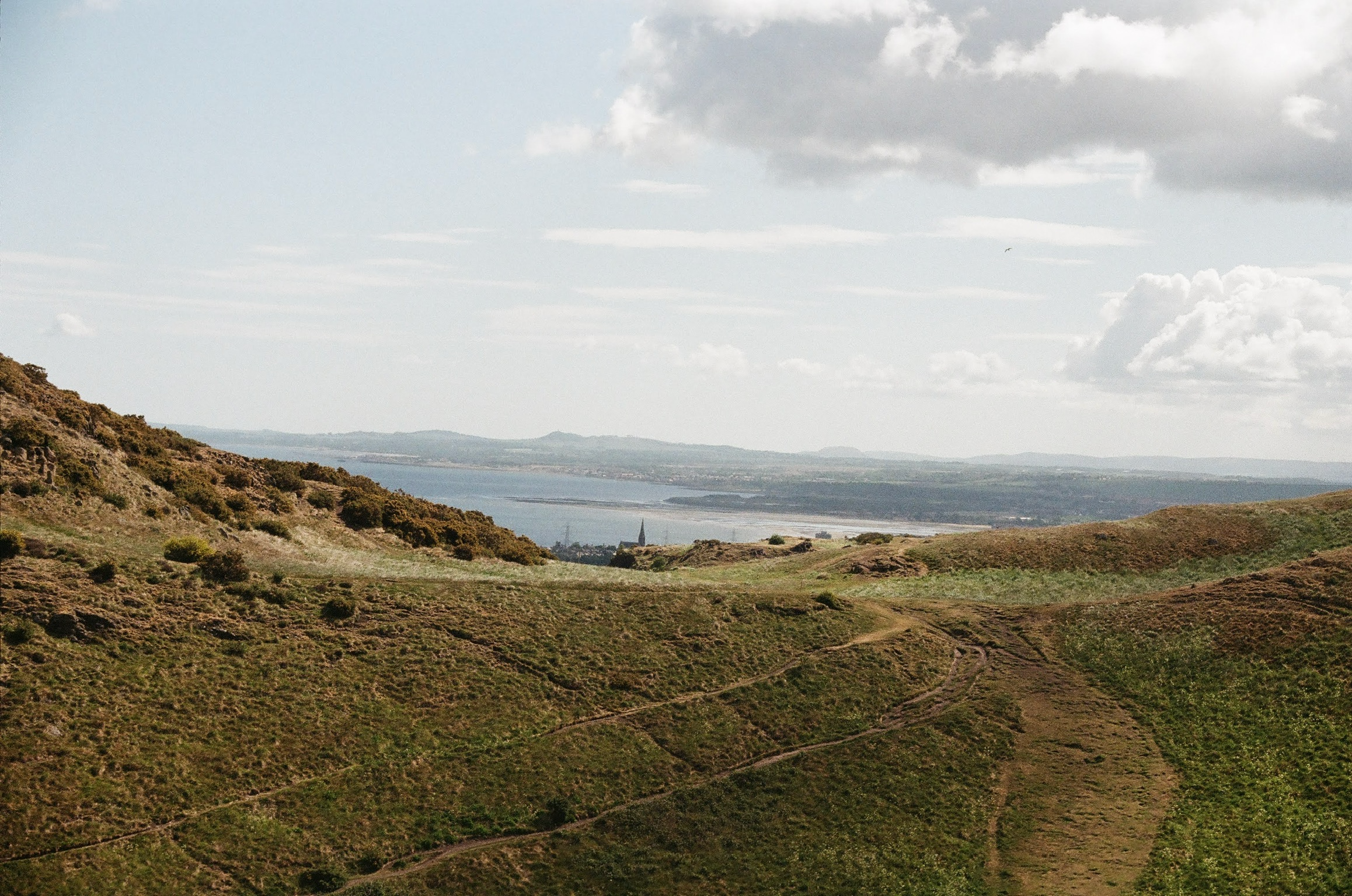 Landscape with rolling green hills, a large body of water in the distance, and a sky with scattered clouds.