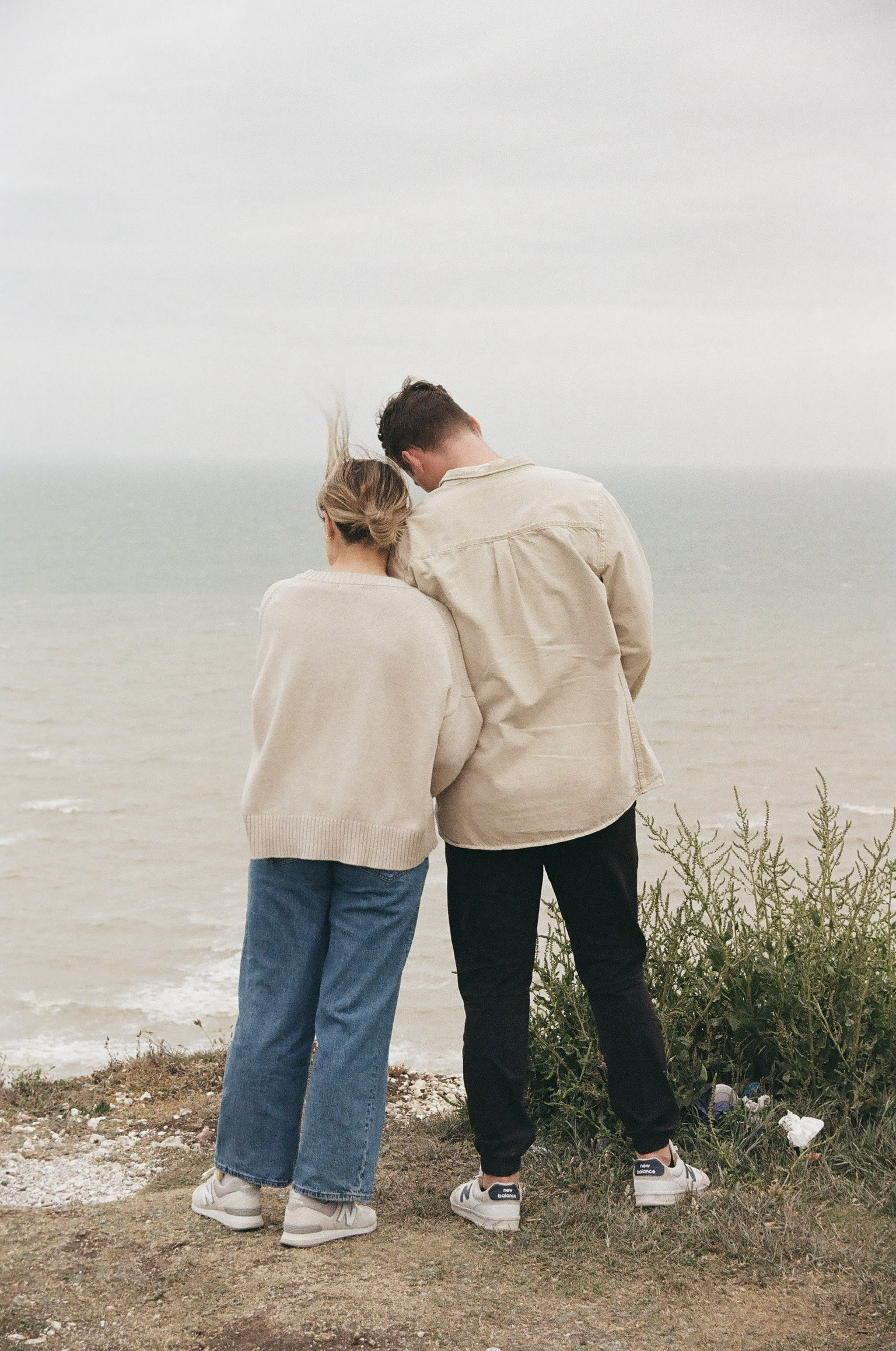 A man and a woman standing on a grassy area by the water, facing away from the camera, with their heads resting against each other, near some shrubs.