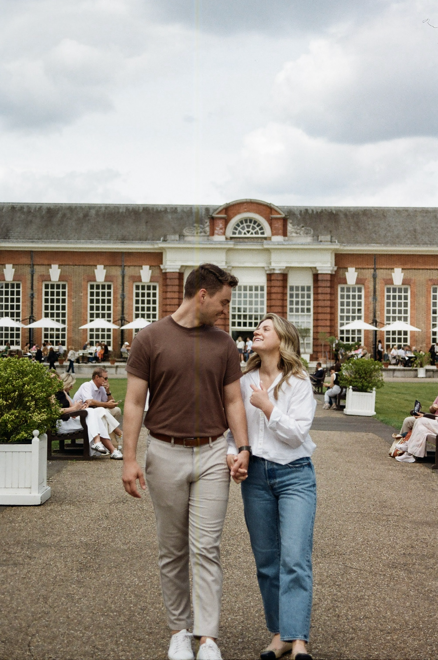 A couple holding hands and smiling at each other walking in a park with a historic building in the background, cloudy sky above.