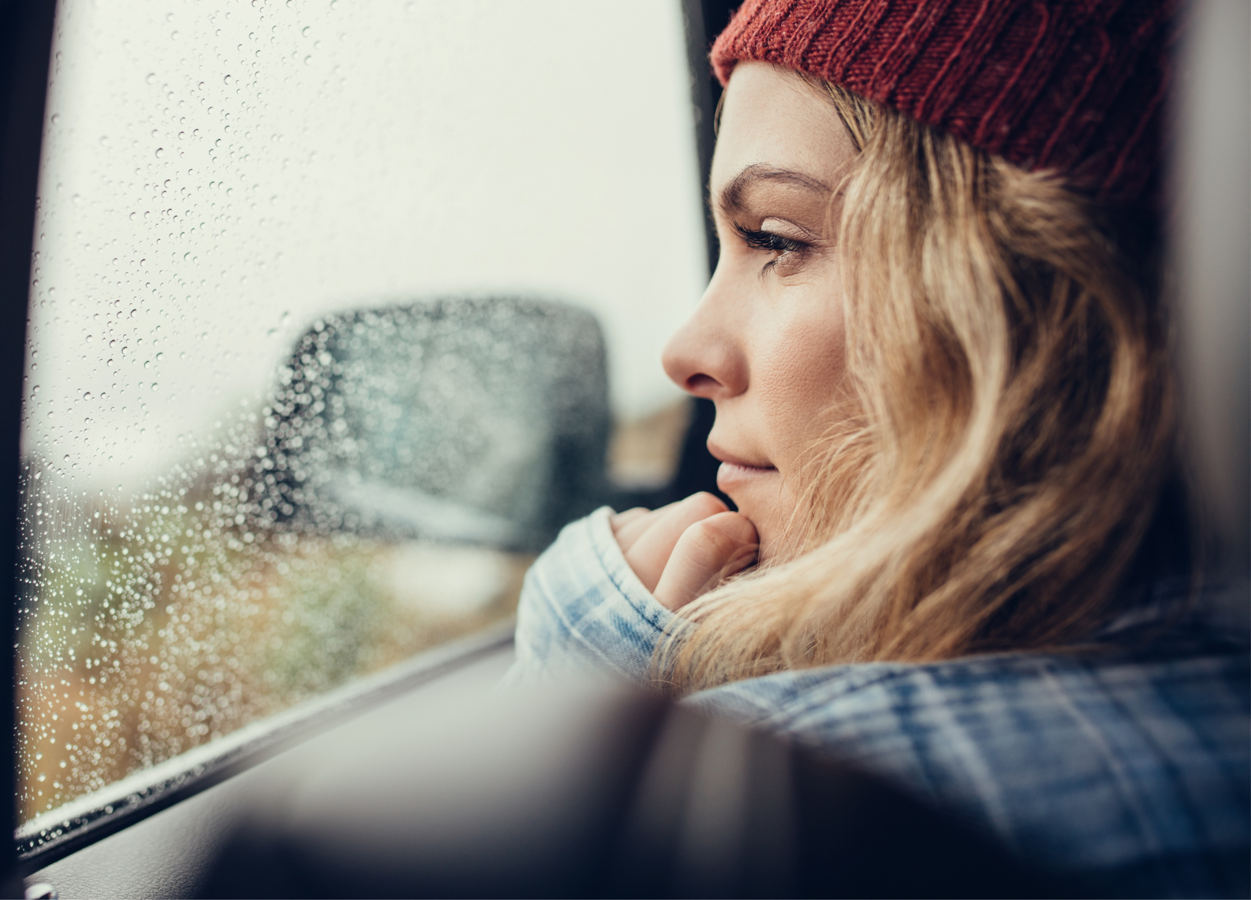 A woman of faith and church leader with curly hair wearing a red beanie, looking thoughtfully out of a car window with raindrops, on a rainy day.