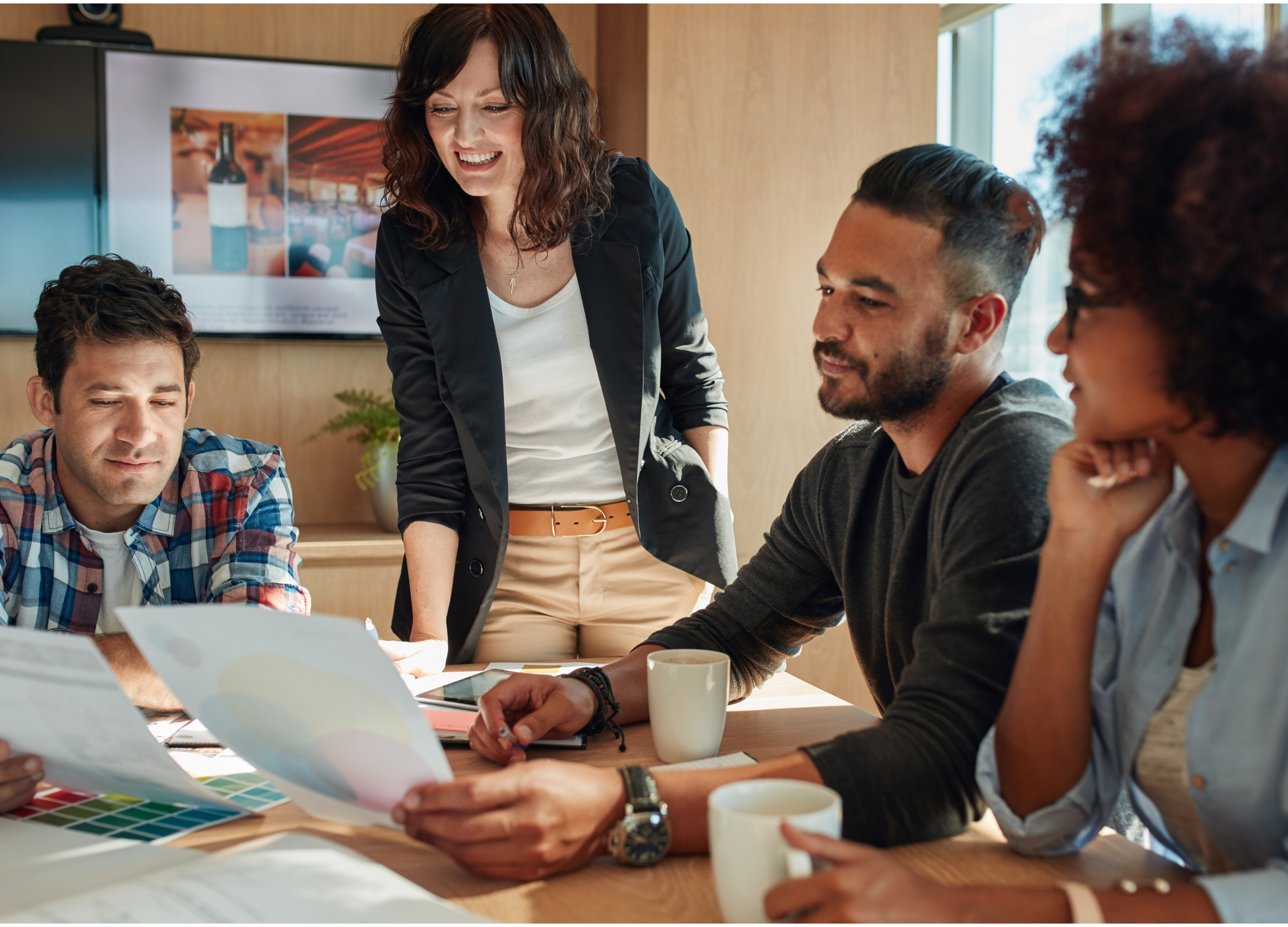 Start-up business team in a meeting room, discussing documents and smiling, with a woman standing and three seated at a table with coffee cups.
