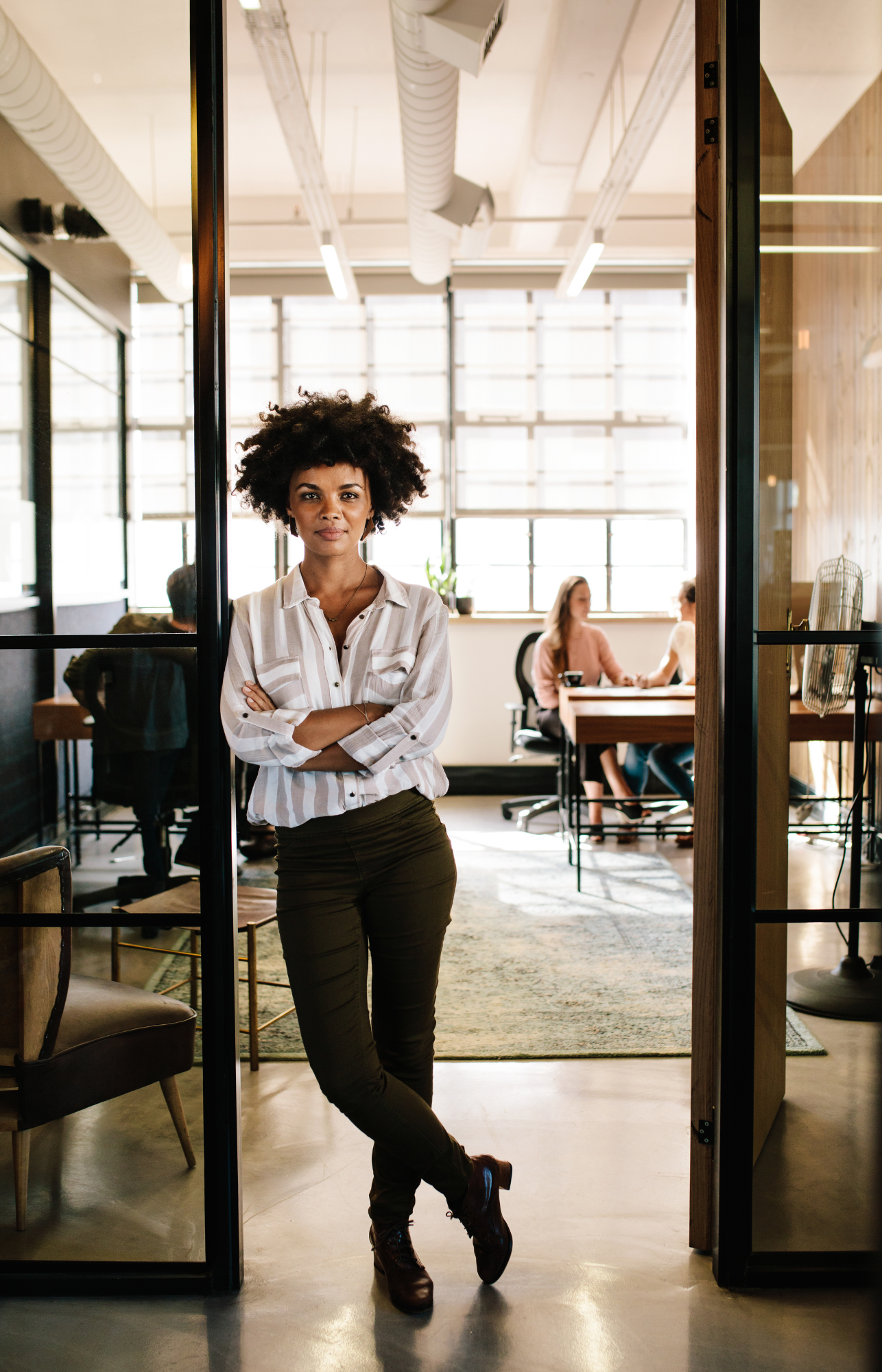 non profit director standing with arms crossed in office, with coworkers working in the background.