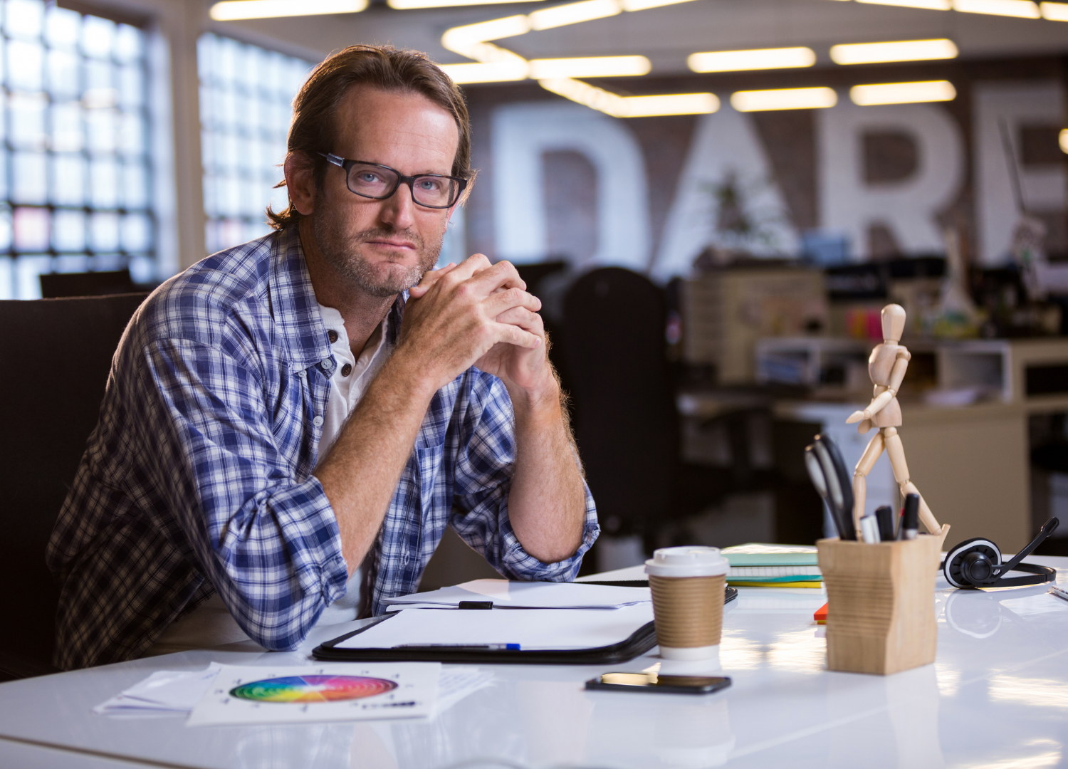 Mission driven entrepreneur with glasses and a beard sitting at a desk in an office, with a cup of coffee, notepad, and pens on the desk, looking at the camera.