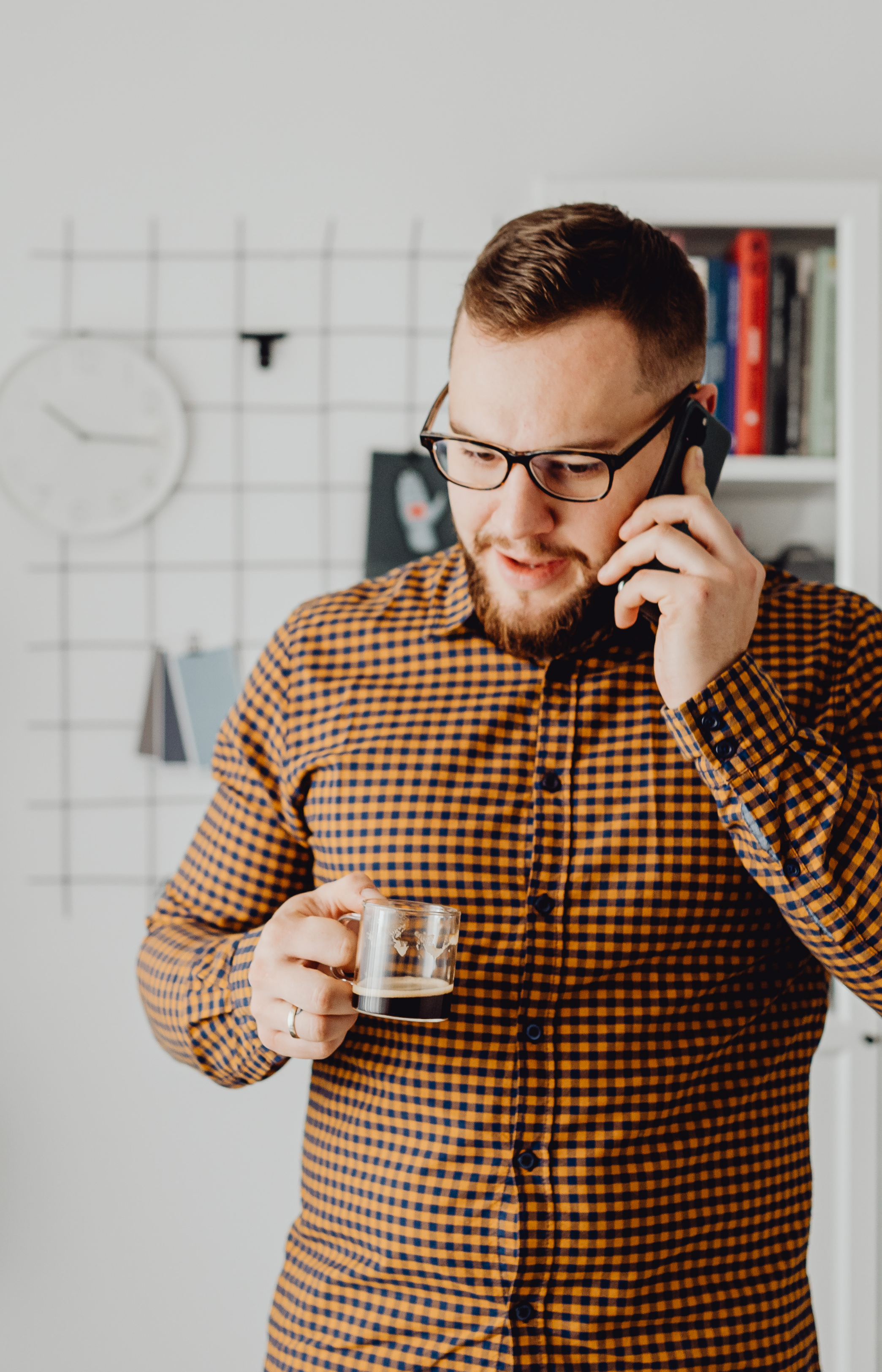 A business owner with glasses and a beard, wearing a checked shirt, talking on a cellphone talking with his start-up coach and holding a small glass of coffee in a modern office or home setting.