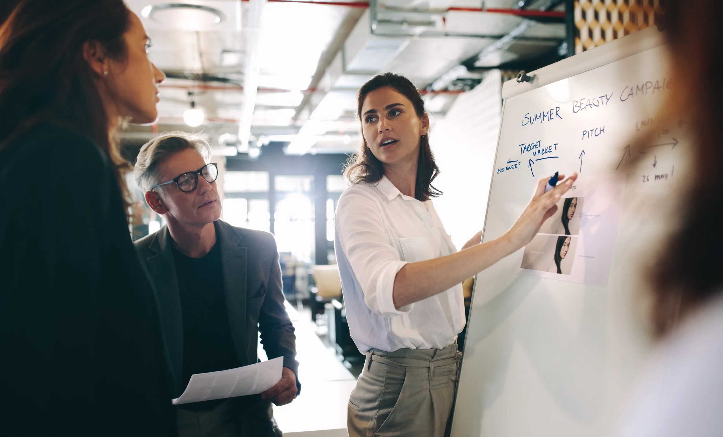 A business consultant presents a marketing plan to a mission driven team on a whiteboard to two colleagues, in an office during a meeting.