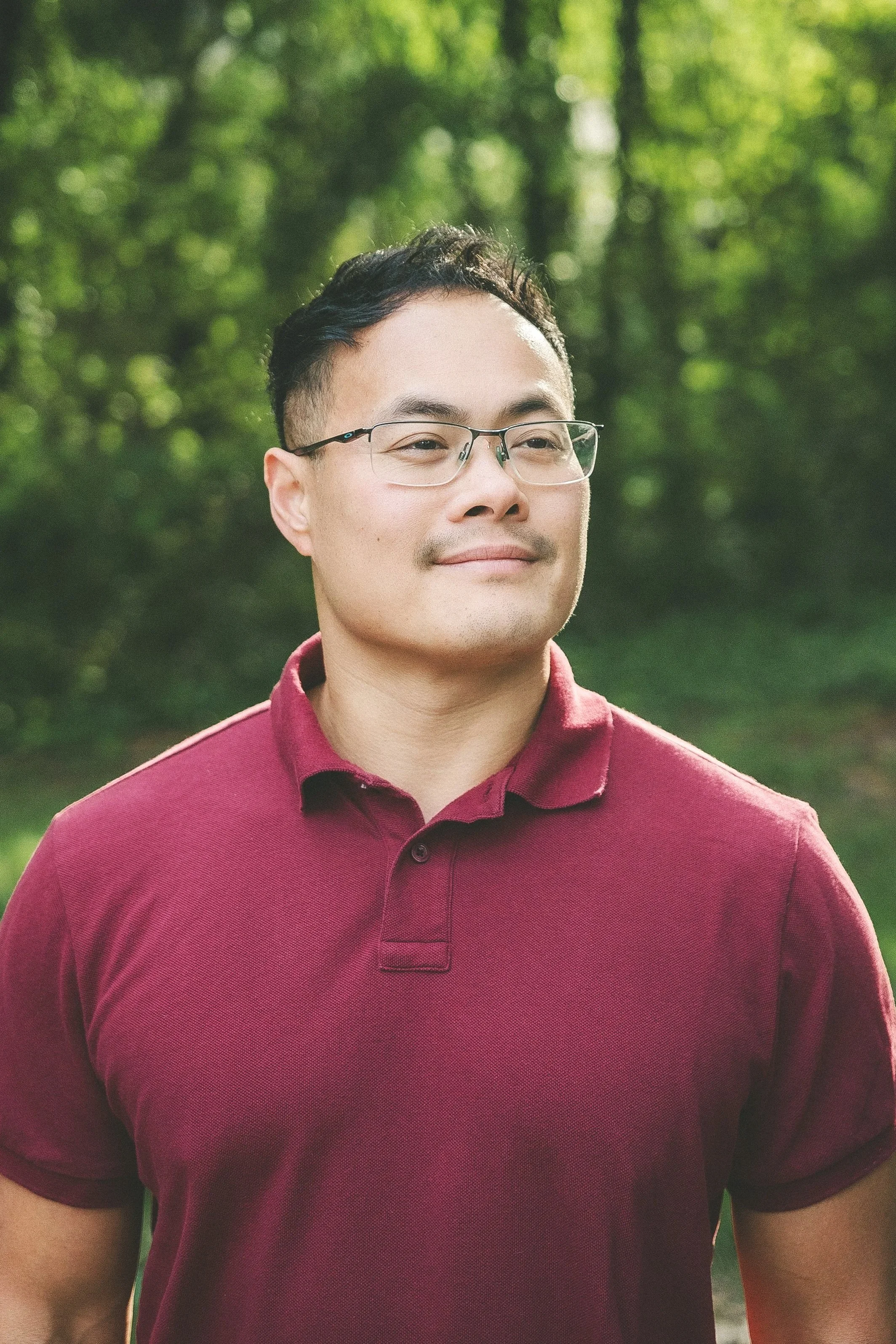 A young man wearing glasses and a maroon polo shirt standing outdoors in a green, wooded area.