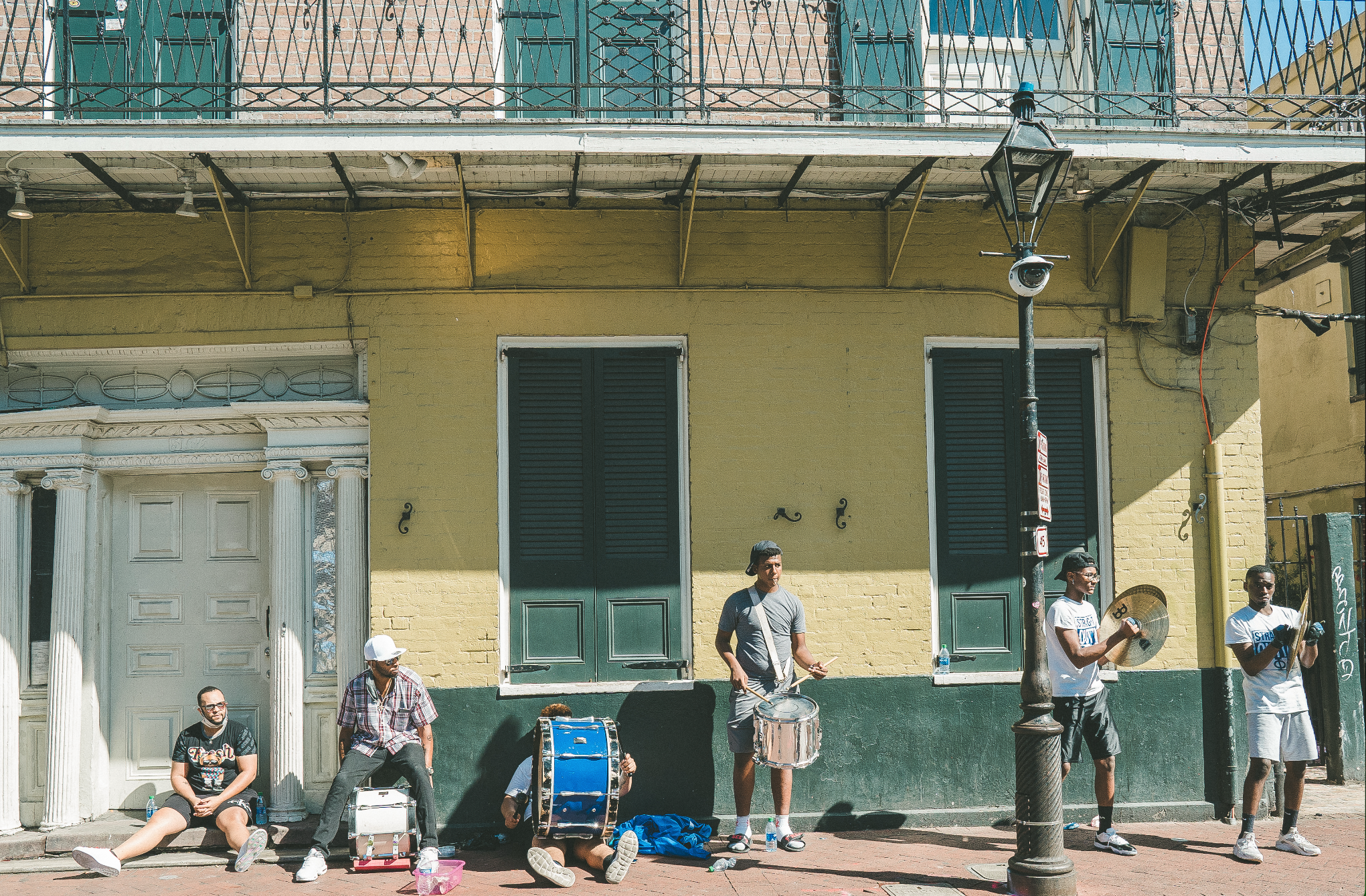Group of street musicians performing on the sidewalk in front of a yellow building with green shutters, with some sitting and others standing and playing instruments.