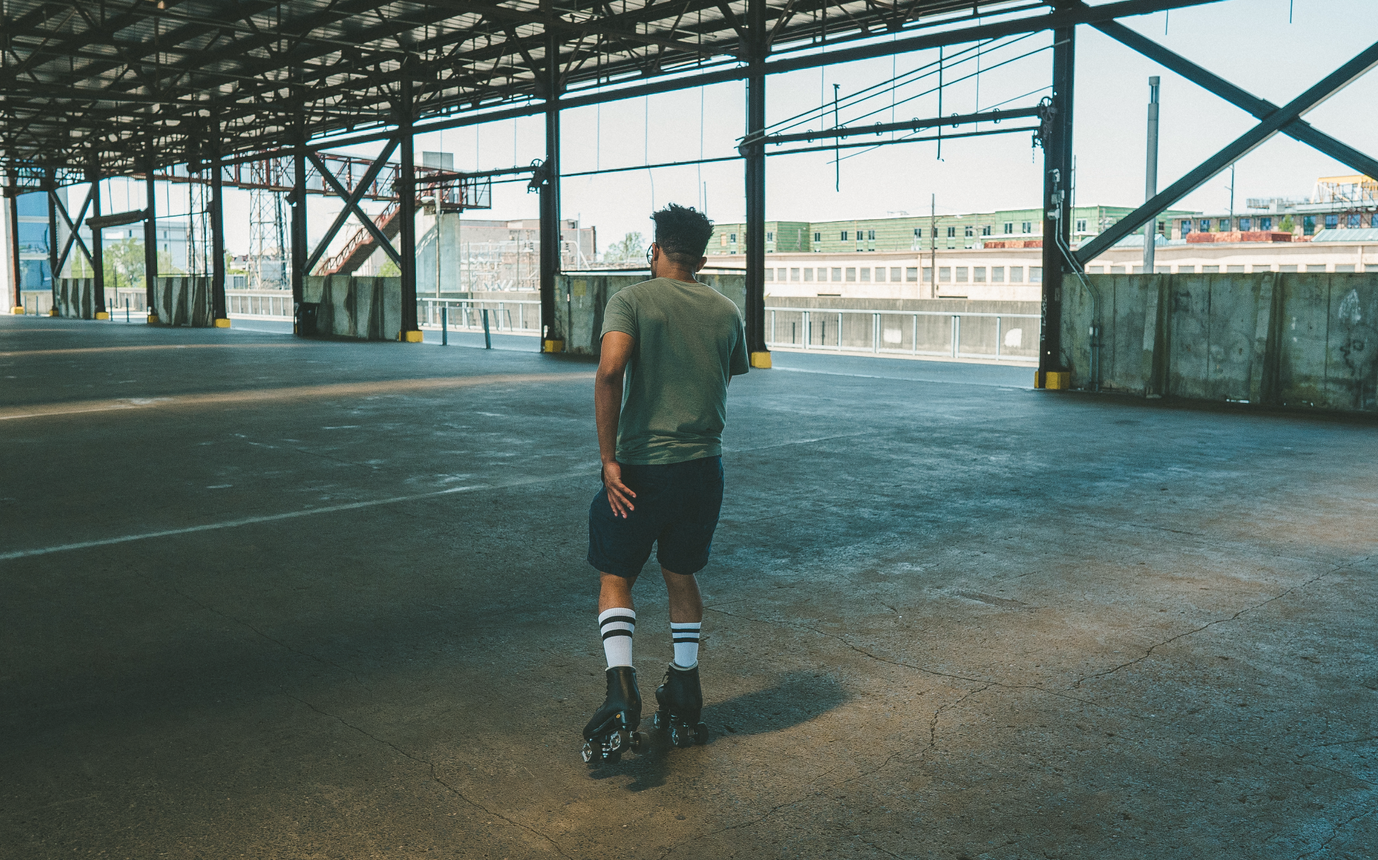 A young man rollerblading in an empty parking garage, wearing a gray t-shirt, black shorts, striped knee-high socks, and black rollerblades.