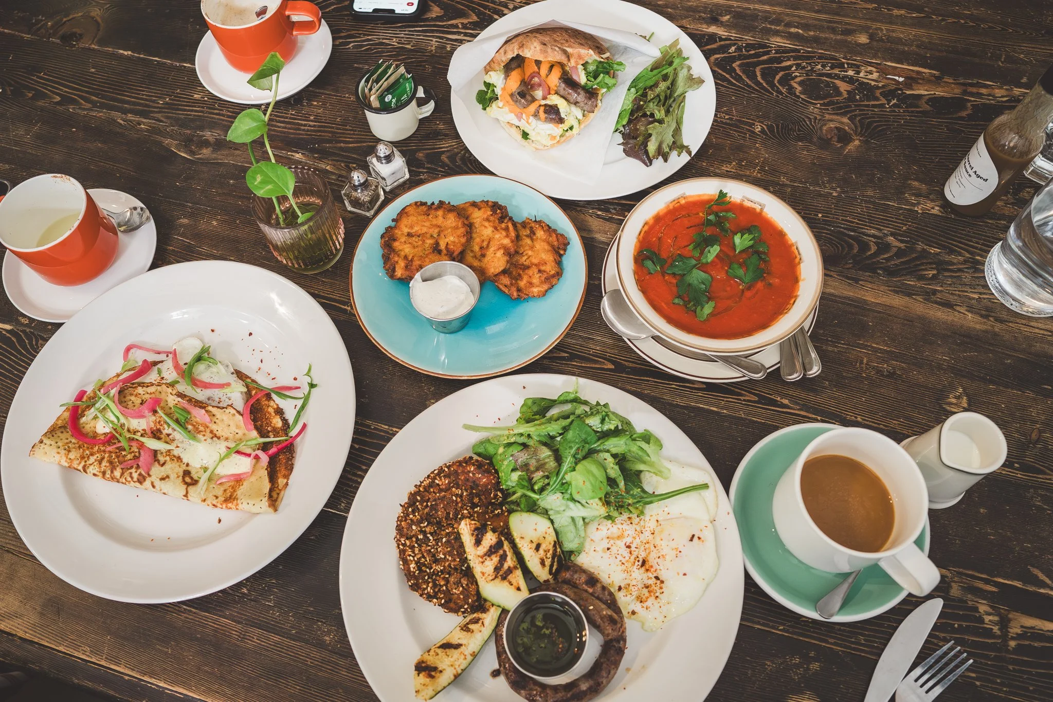 A variety of dishes on a wooden table including a slice of pizza, a salad with fried eggplant, a bowl of tomato soup garnished with herbs, a sandwich with vegetables and meat, and roasted eggplant with vegetables, along with cups of coffee, water, and tea.