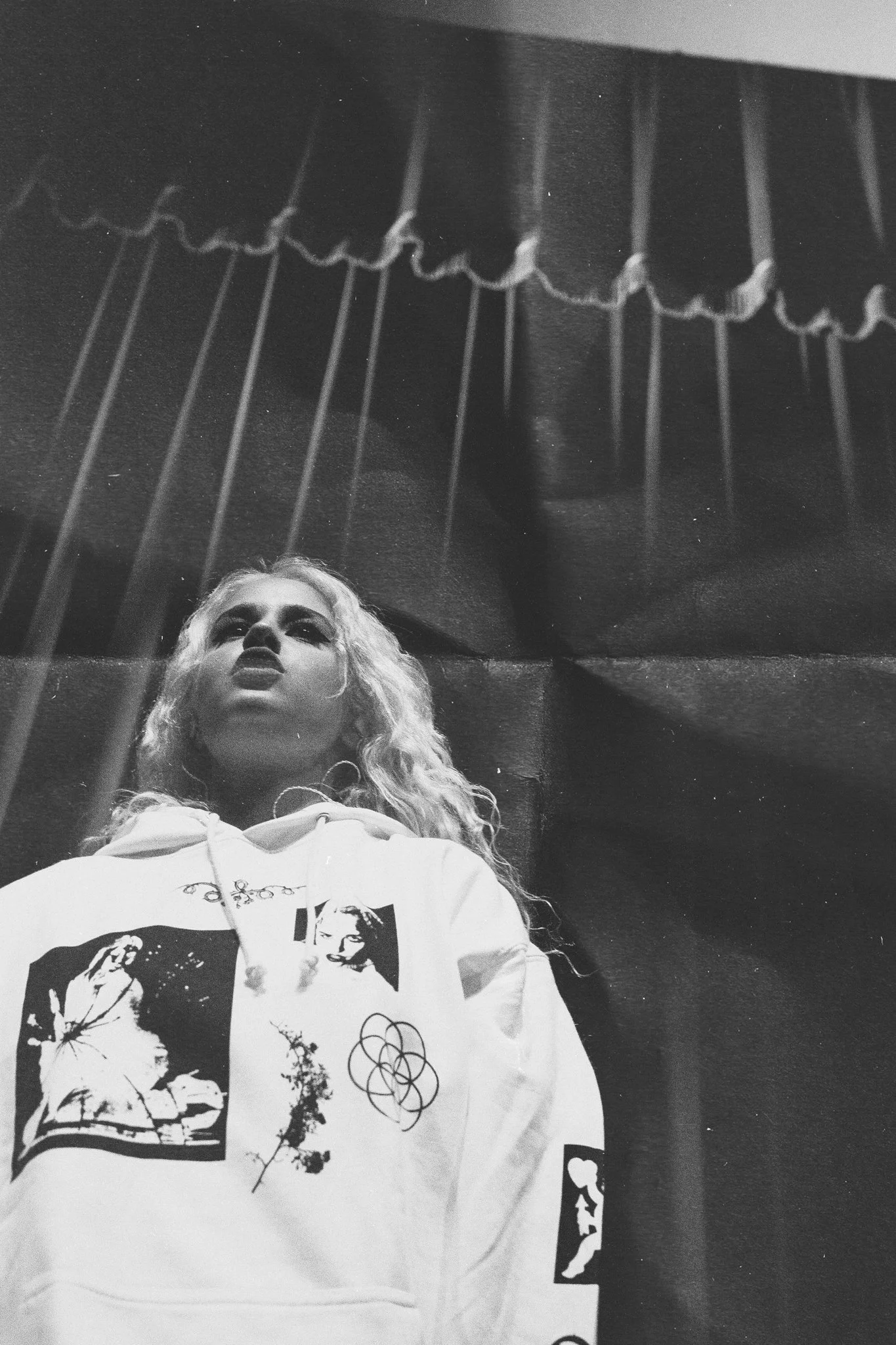 A woman with curly blonde hair looking up, wearing a white hoodie with various black graphic designs, in a room with a dark background and ceiling decoration.