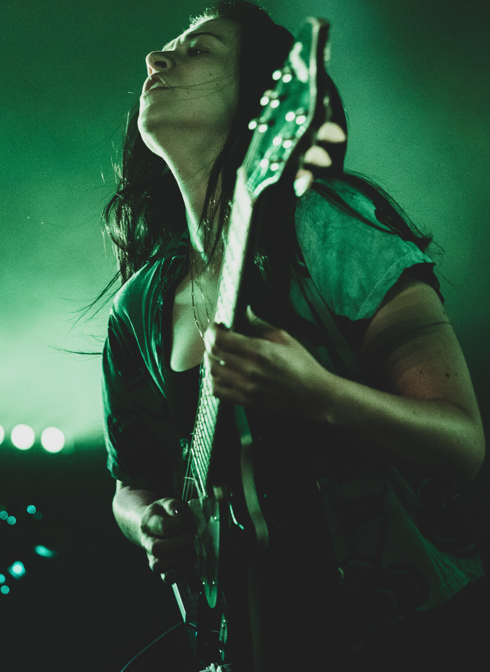 A woman with dark hair playing an electric guitar on stage, with green stage lighting.
