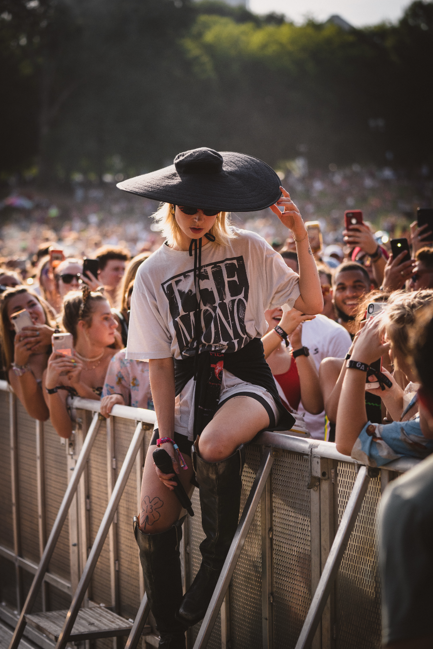 A woman with blonde hair wearing large black hat, sunglasses, oversized t-shirt, and thigh-high boots leaning on a barrier at an outdoor concert with a crowd taking photos behind her.