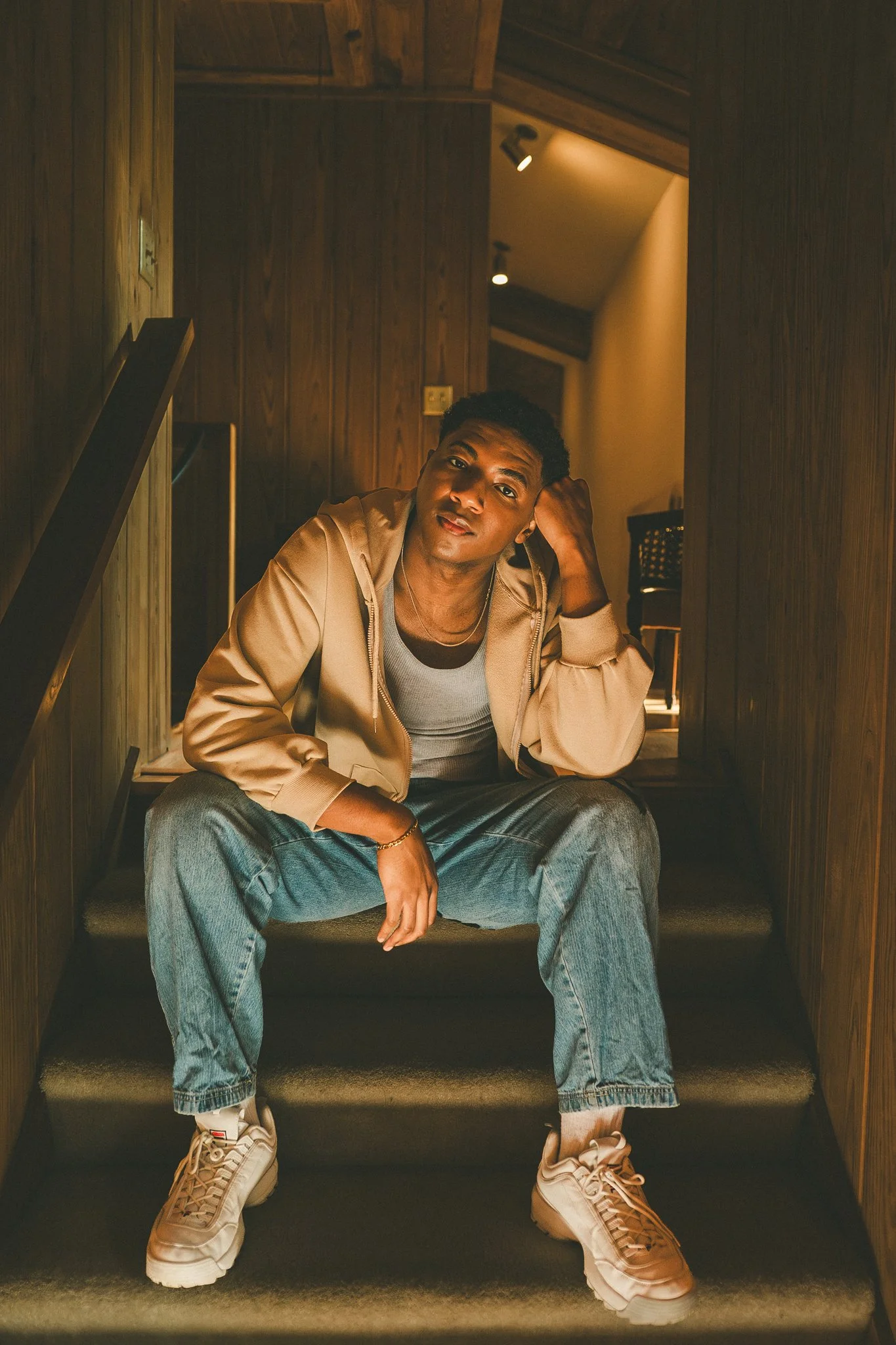 A young man sitting on stairs inside a wooden-paneled room, with one arm resting on his knee and the other hand behind his head, looking at the camera.