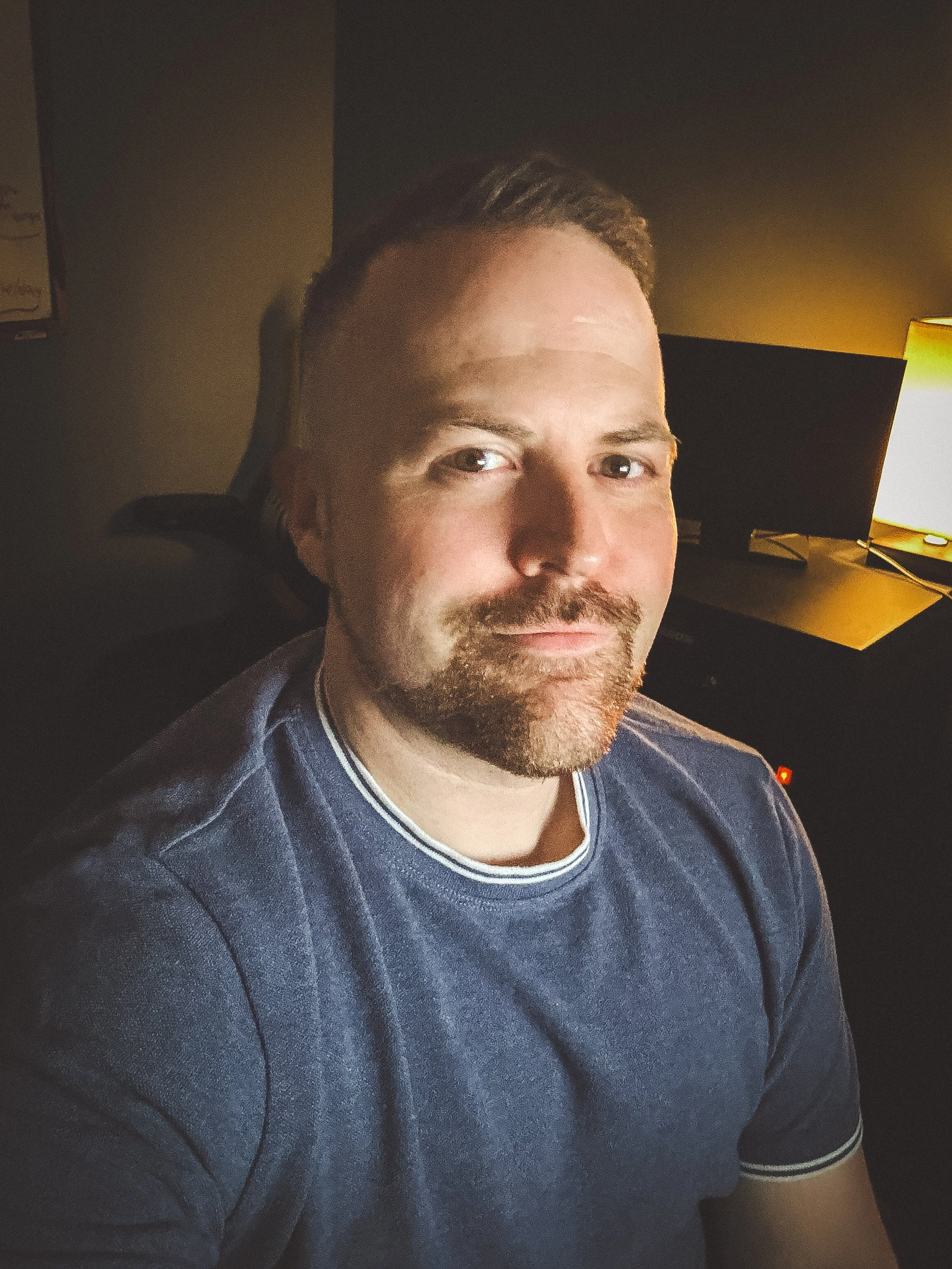 A man with short brown hair and a beard, wearing a blue t-shirt, taking a selfie in a dimly lit room with a computer monitor and a lamp in the background.