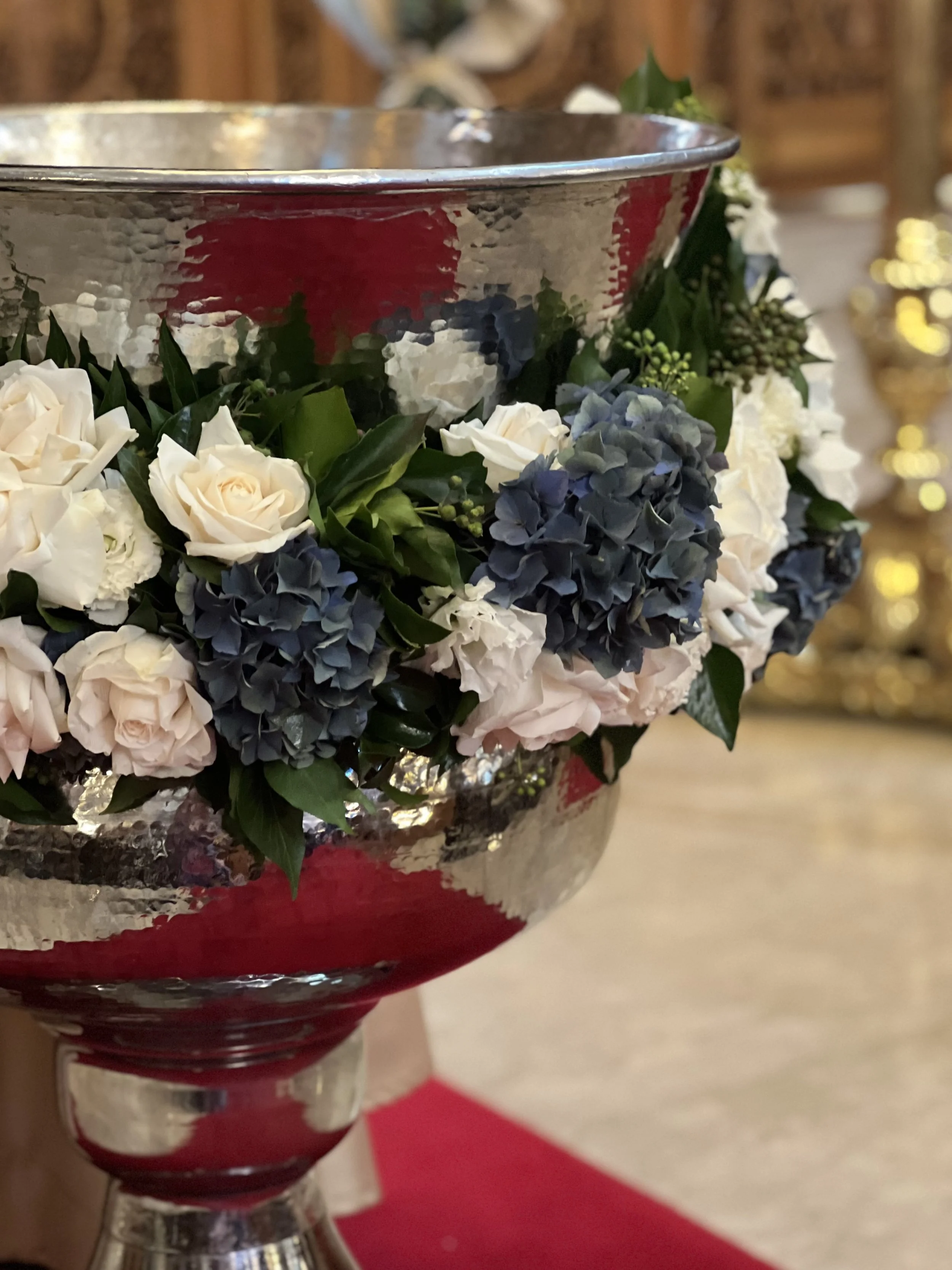 Close-up of a silver trophy cup decorated with white roses, blue hydrangeas, and greenery, placed on a table with a red tablecloth, with a blurred background.