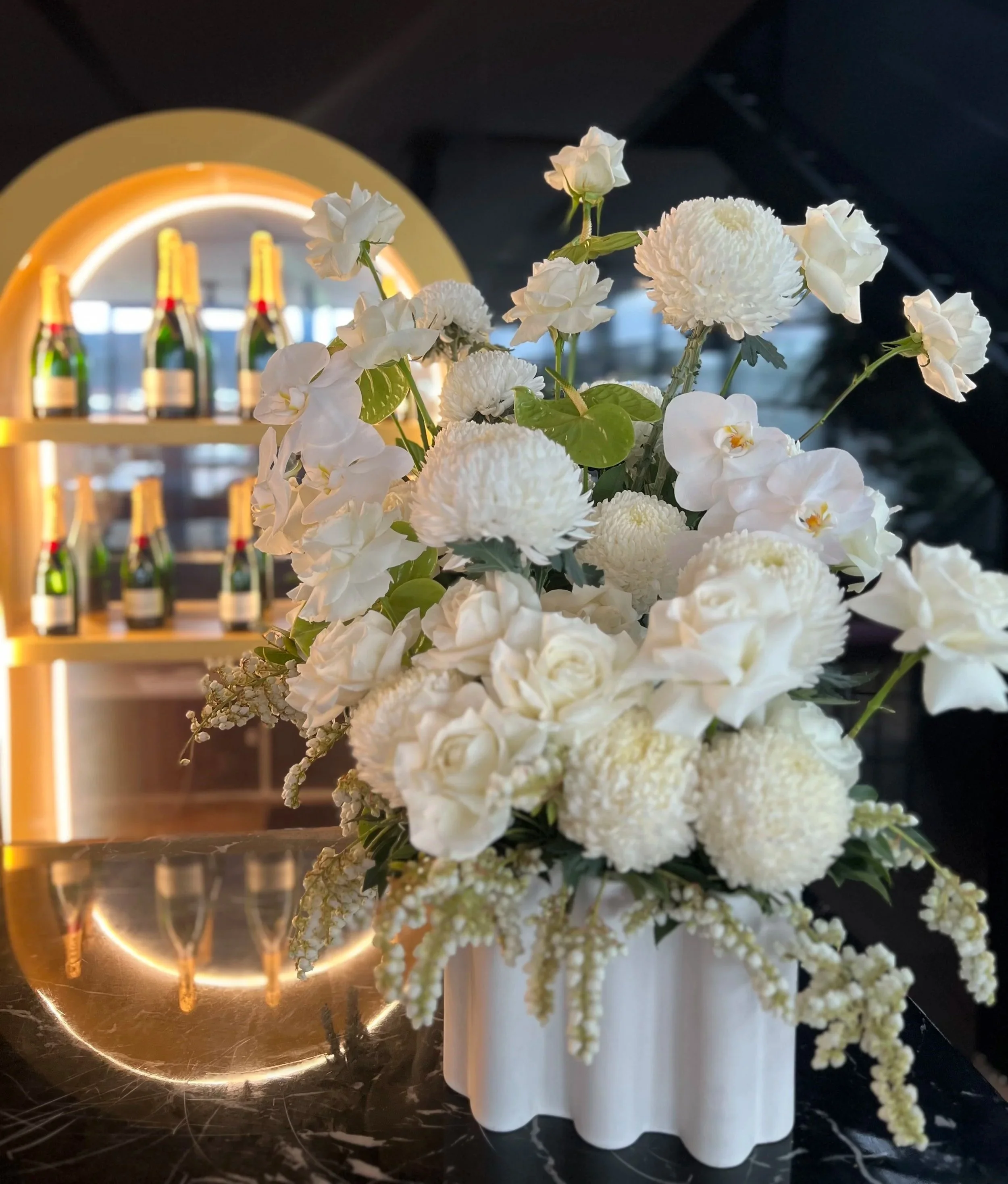 A floral centerpiece with white flowers, including roses, carnations, and orchids, in a white vase on a marble surface. In the background, there is a bar shelf with bottles and a glowing circular lighting feature.