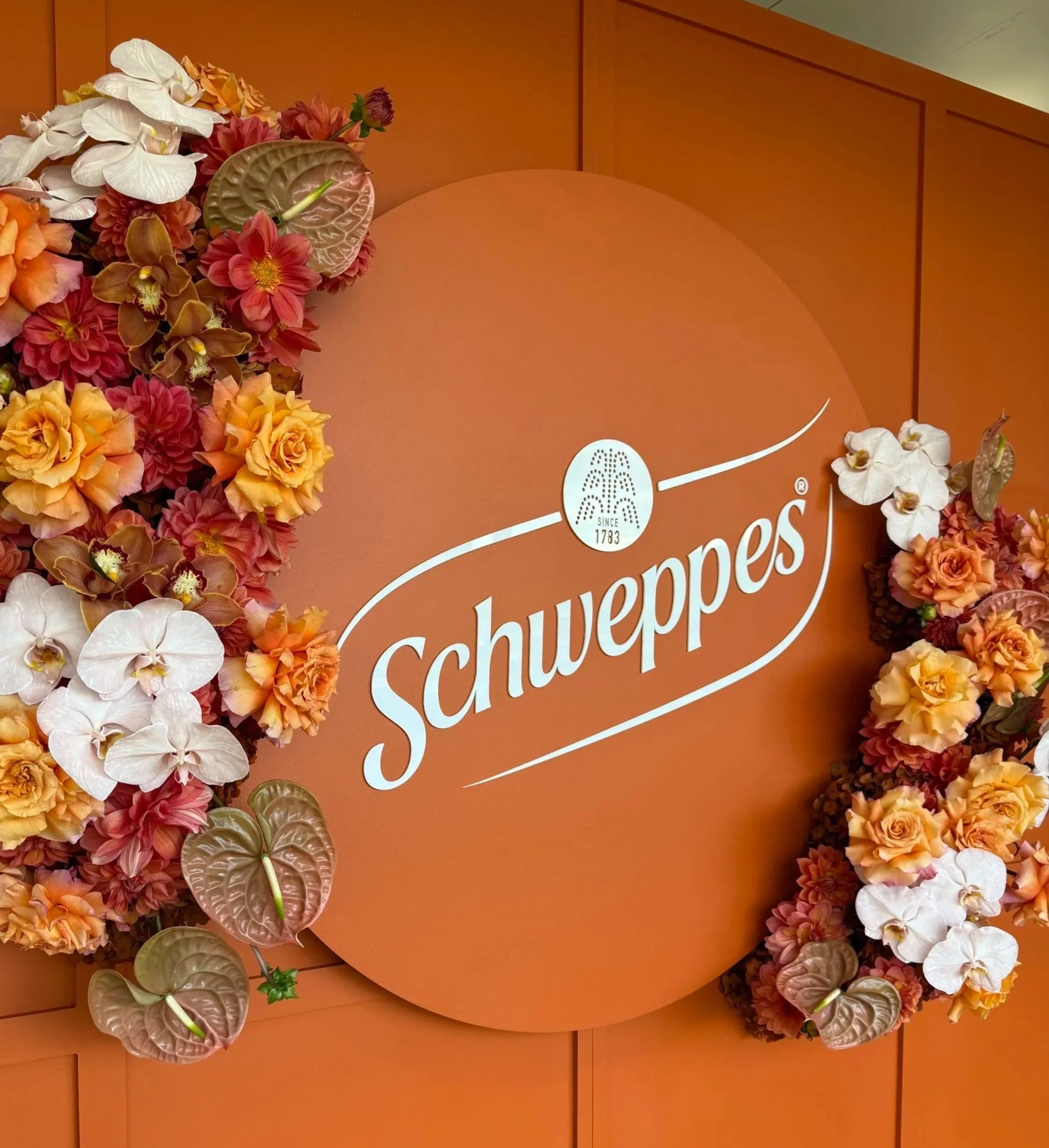 A floral arrangement with pink, white, and orange flowers arranged around an orange background with partial text that includes the word 'Schweppes' and the phrase 'Since 1783'.