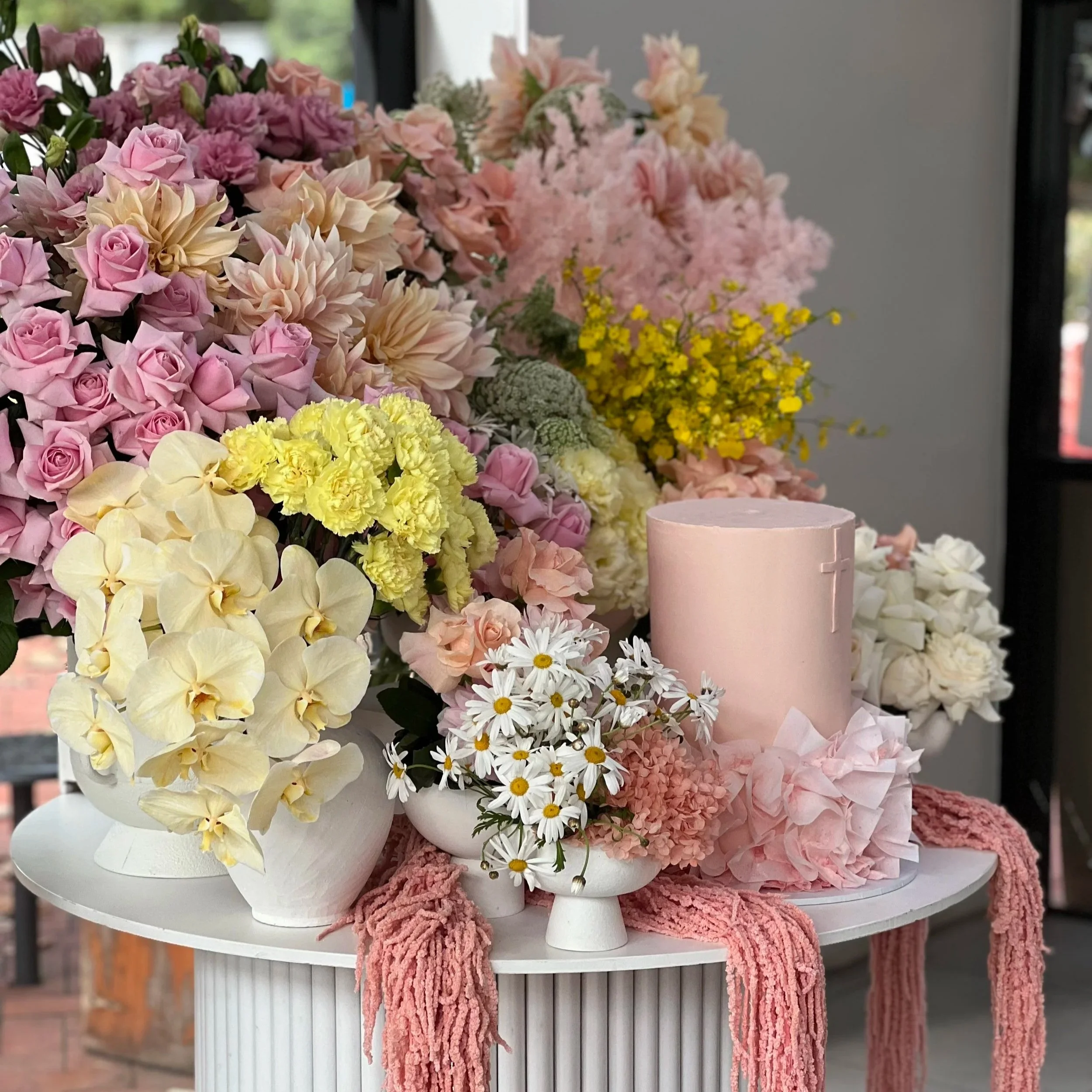 Colorful floral arrangement including pink roses, cream-colored flowers, pink carnations, white daisies with yellow centers, and other flowers in white vases on a round white table.