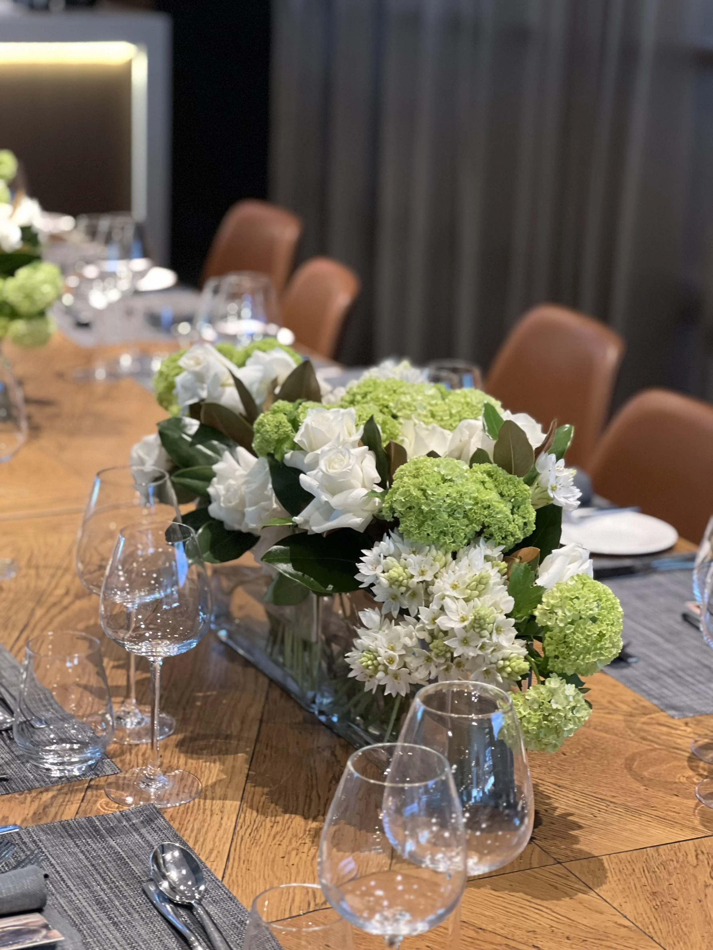 A table decorated with a floral centerpiece of white and green flowers, set with glassware and silverware, and surrounded by brown chairs.