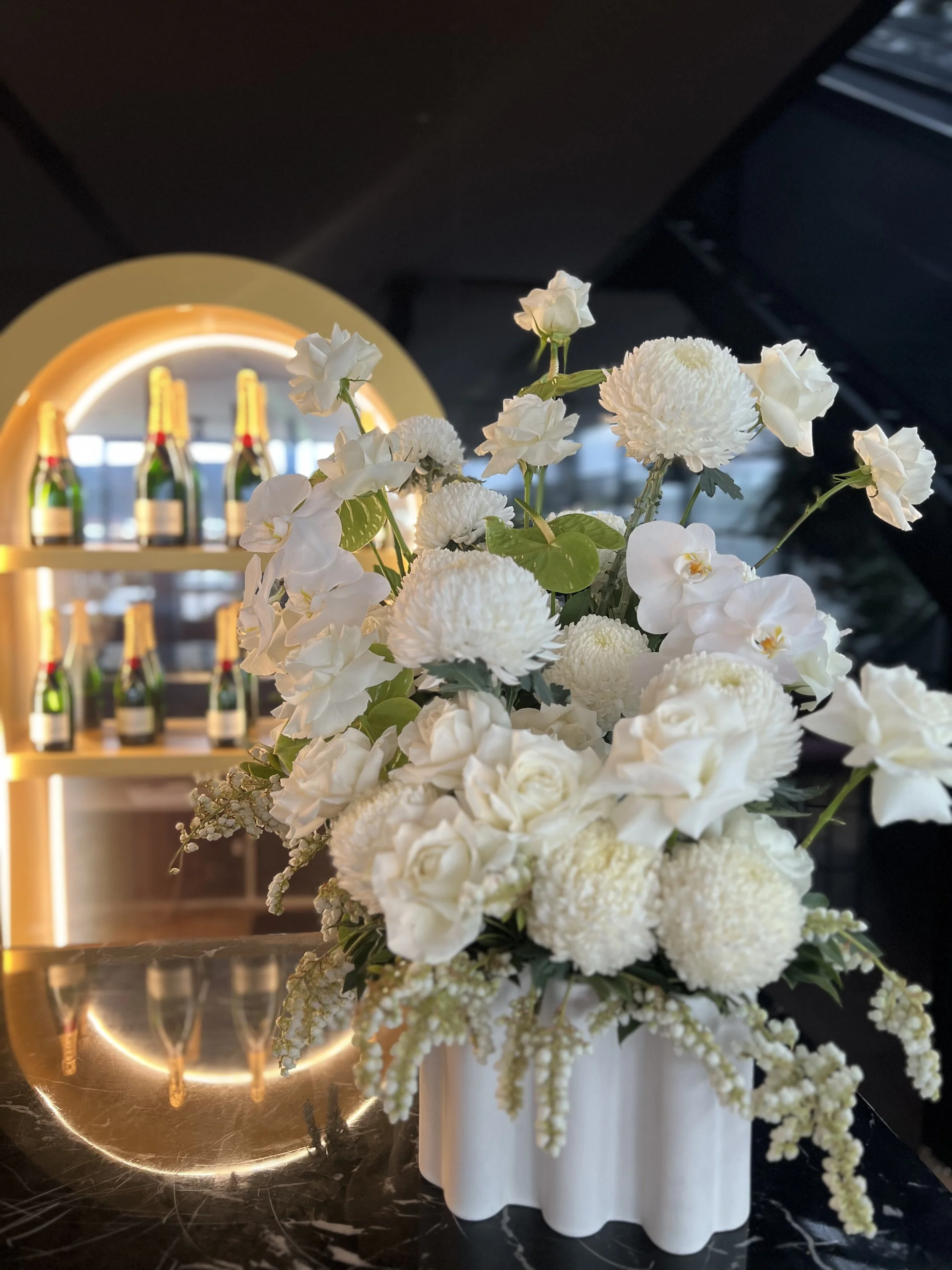 A bouquet of white flowers, including roses, hydrangeas, and orchids, in a white vase, placed on a dark marble surface. In the background, a wooden bar shelf with bottles is illuminated by warm lighting.